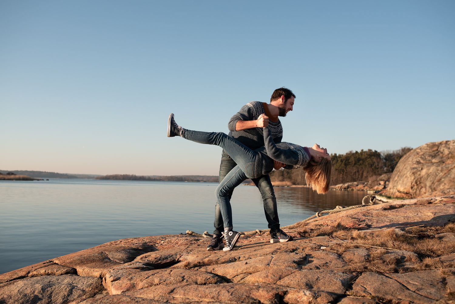 Playful couple performing dramatic dip pose on rocky shoreline at sunset.