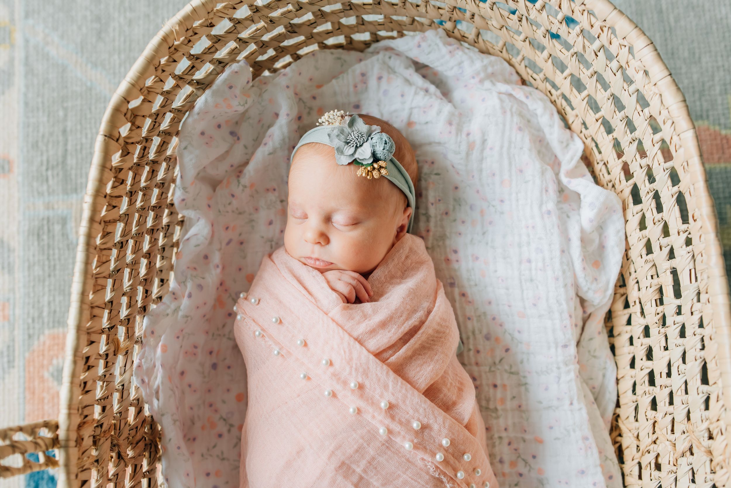 A newborn baby is swaddled in a pink wrap and a flower headband in a Moses basket for a photo session in Fair Haven NJ.
