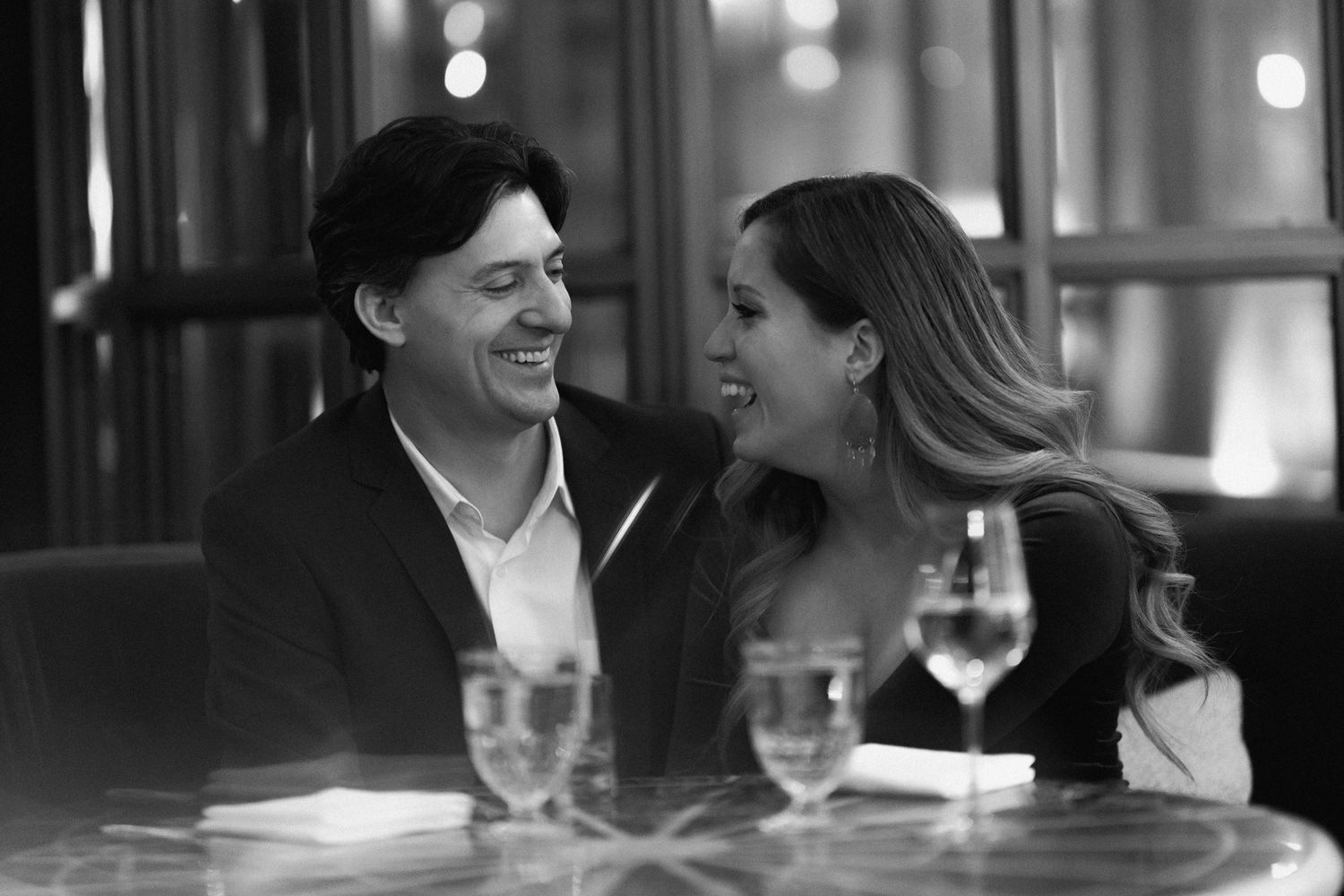 A romantic black and white candid moment of a couple sharing laughter at a restaurant table with wine glasses.
