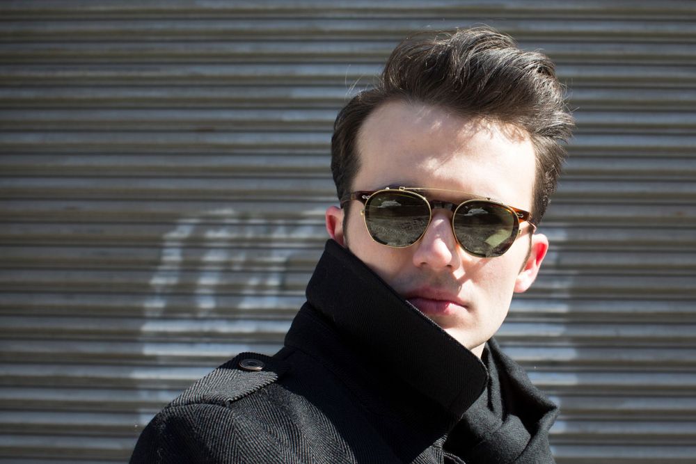 Portrait shot of person in sunglasses and black coat against industrial backdrop.
