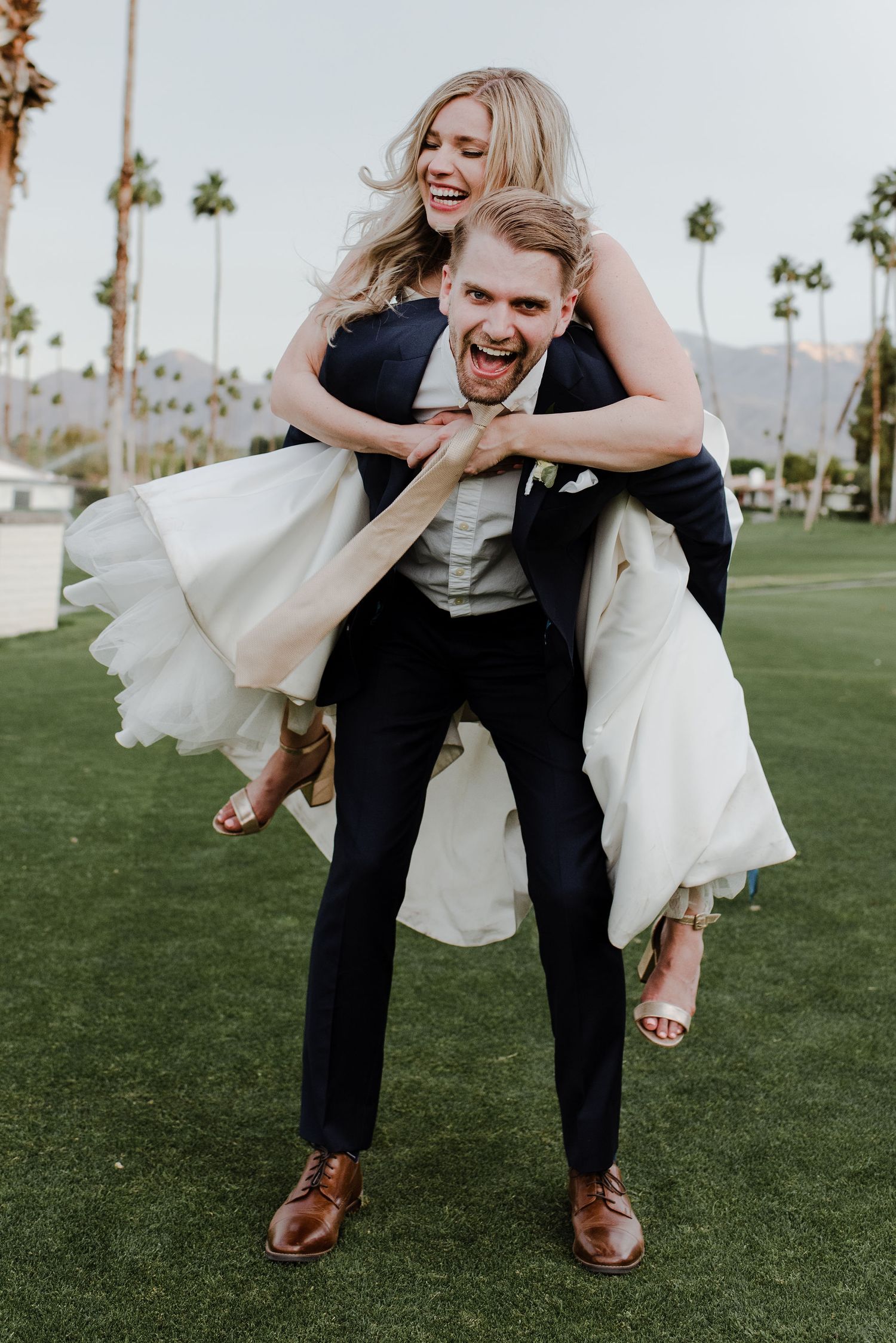 Bride and groom laugh facing camera while he gives her a piggy-back in her bridal gown on a palm tree covered golf green