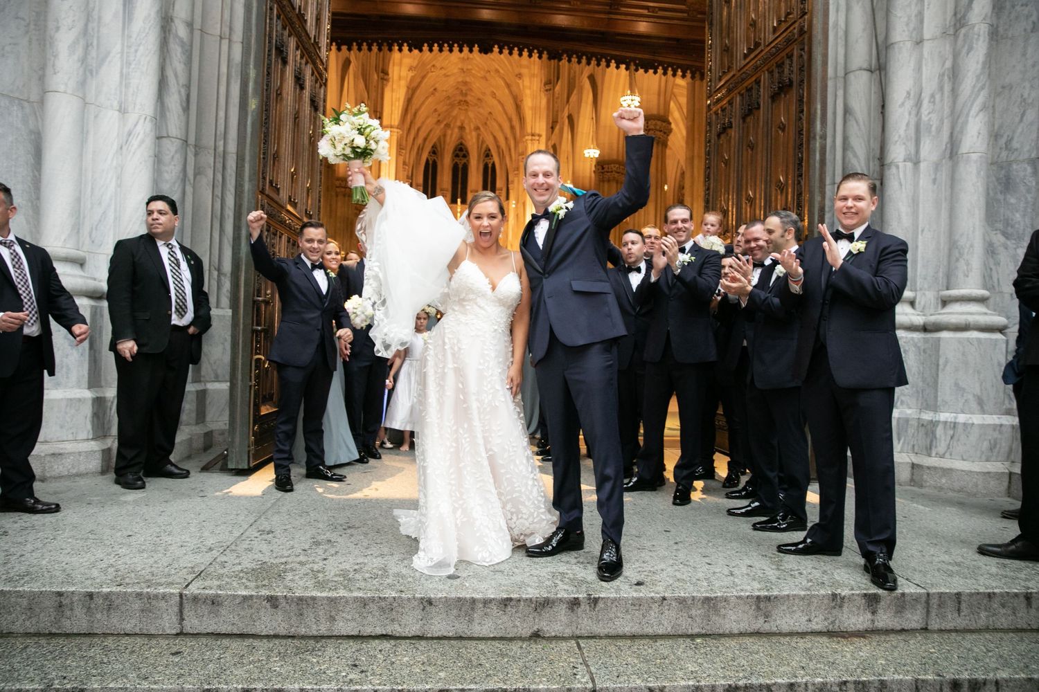 Wedding party celebrating on church steps as guests applaud the newlyweds.