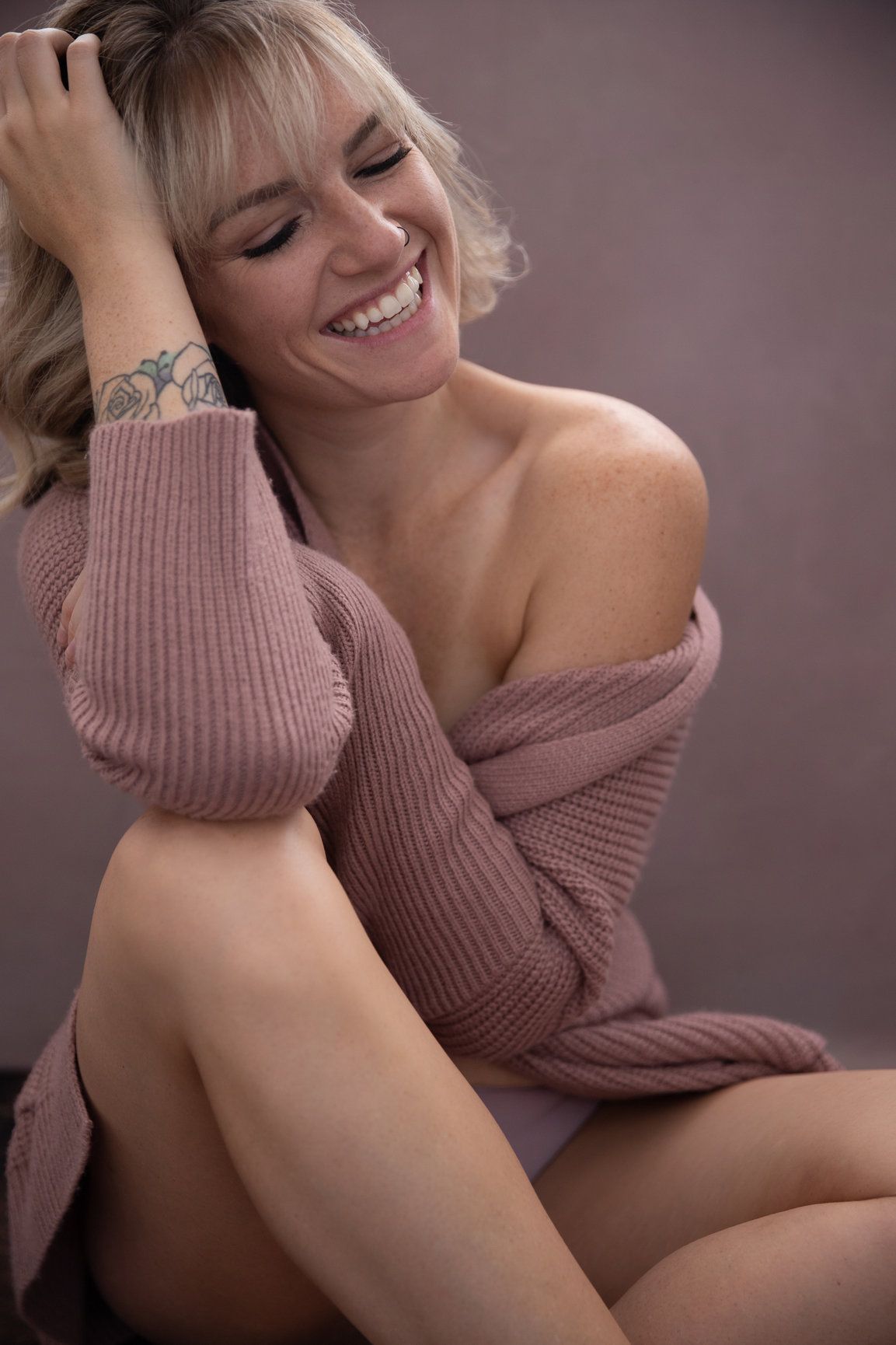 Intimate boudoir portrait showing joyful expression and cozy mauve sweater in soft lighting.