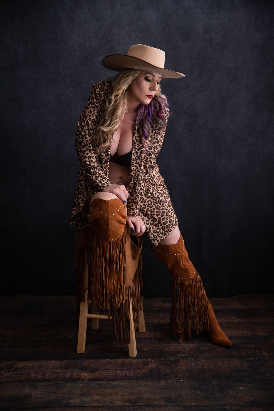 Fashion portrait with western boots, leopard print coat and wide brim hat against dark backdrop.