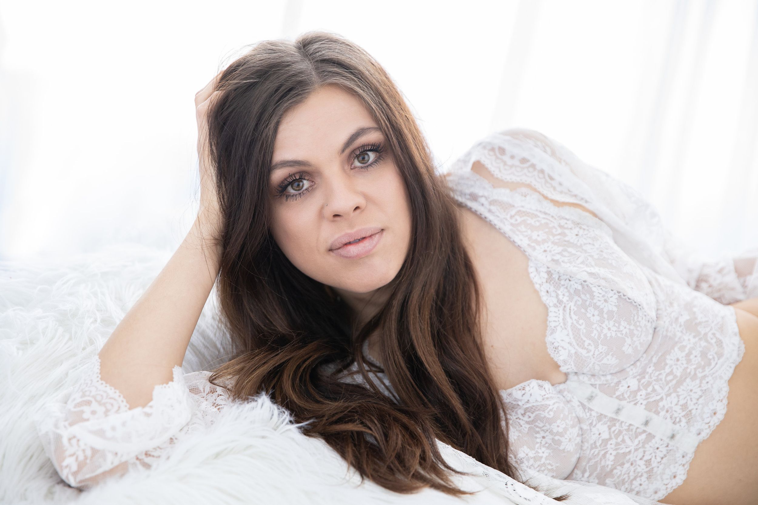 Woman with long dark hair lying on white bedding wearing lacy lingerie top poses sensually.