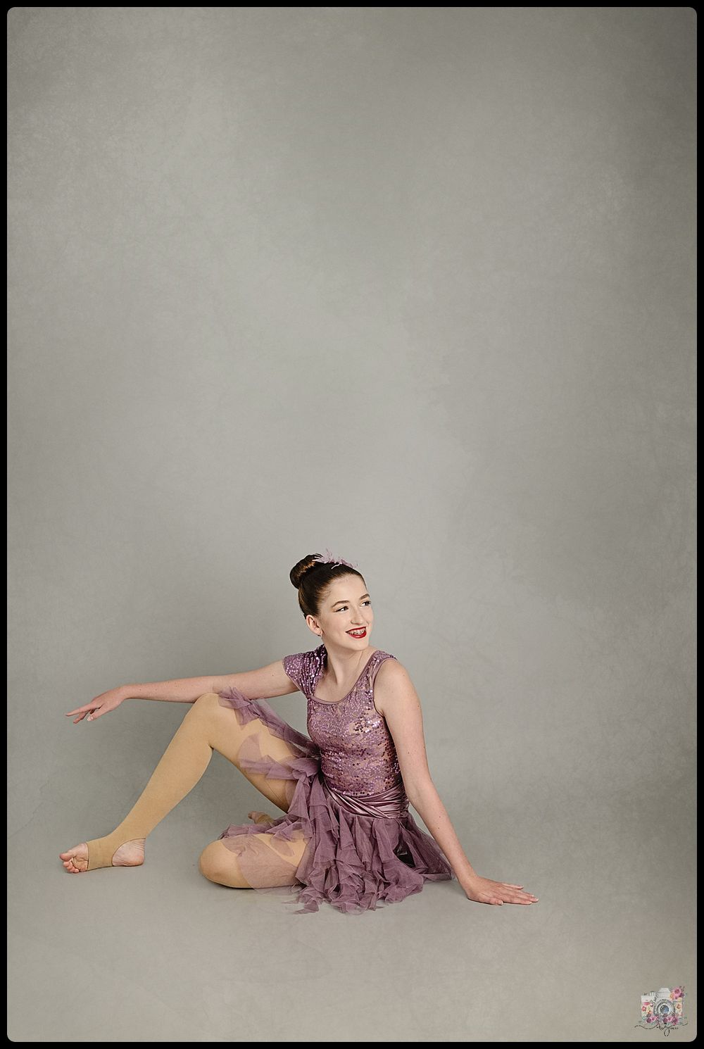 A ballerina in a purple dress sits gracefully on the floor in a studio setting.