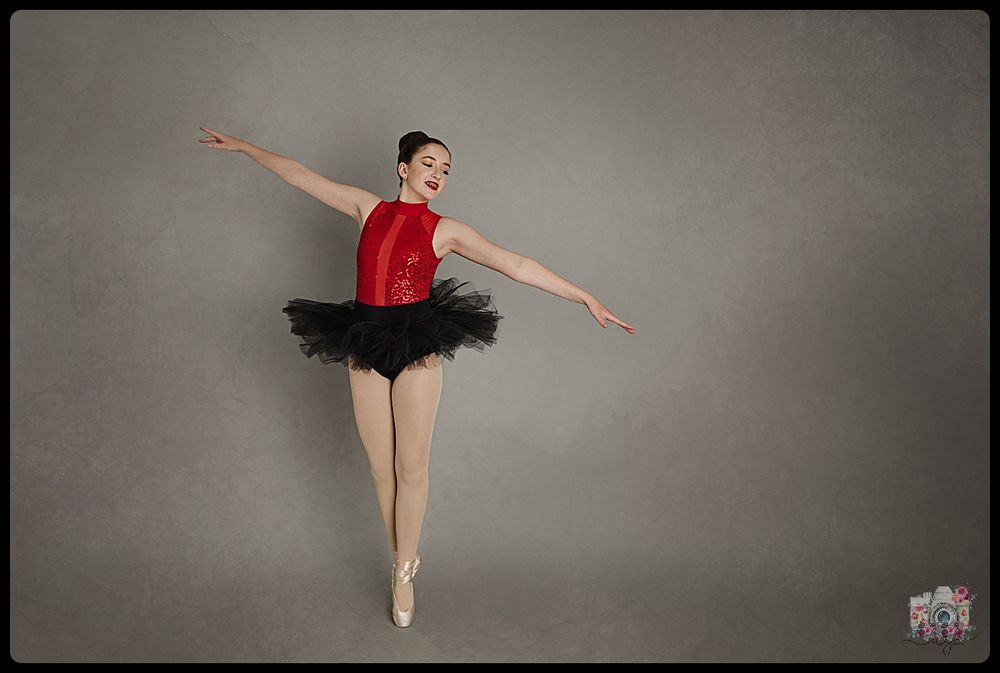 Ballet dancer in red leotard and black tutu performs an arabesque pose against grey backdrop.