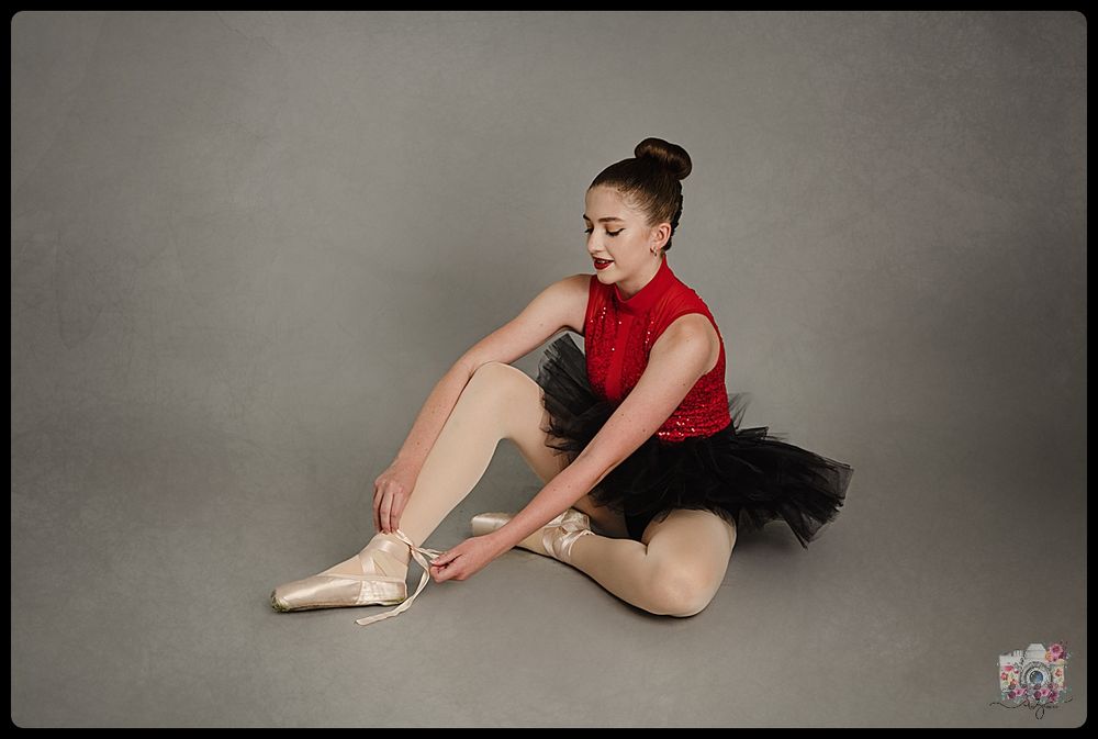 Dancer in red top and black tutu adjusts pointe shoes while seated on floor.