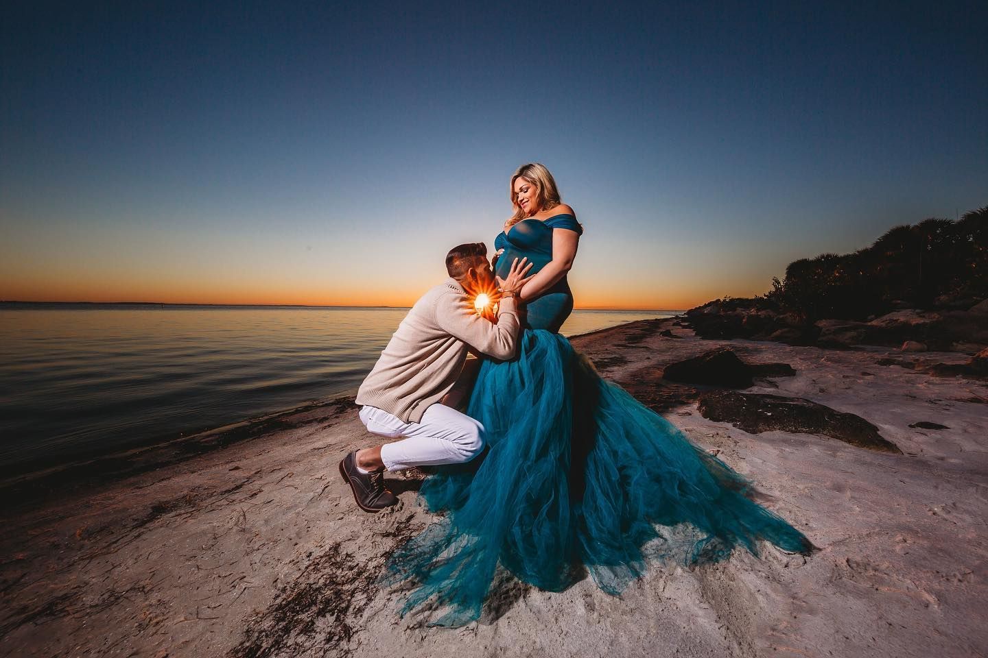Dramatic sunset maternity photo with flowing blue dress on rocky coastline.