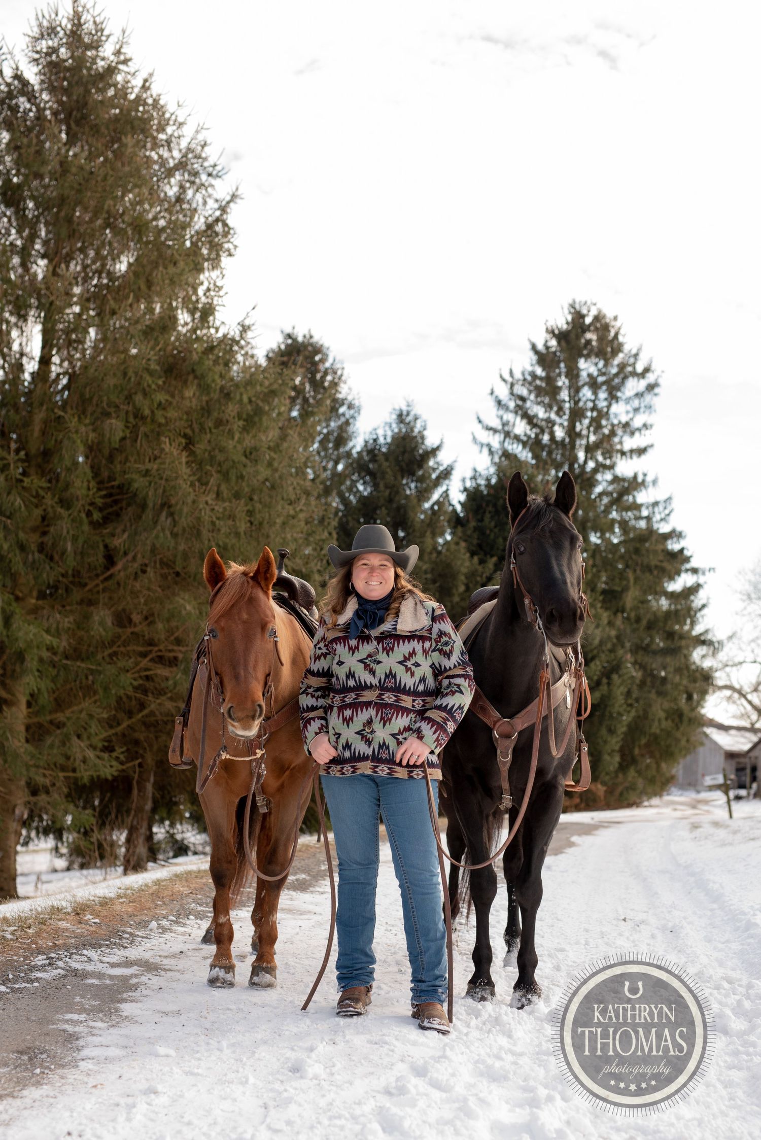 Two horses, one chestnut and one black, stand with their handler on a snowy winter path.