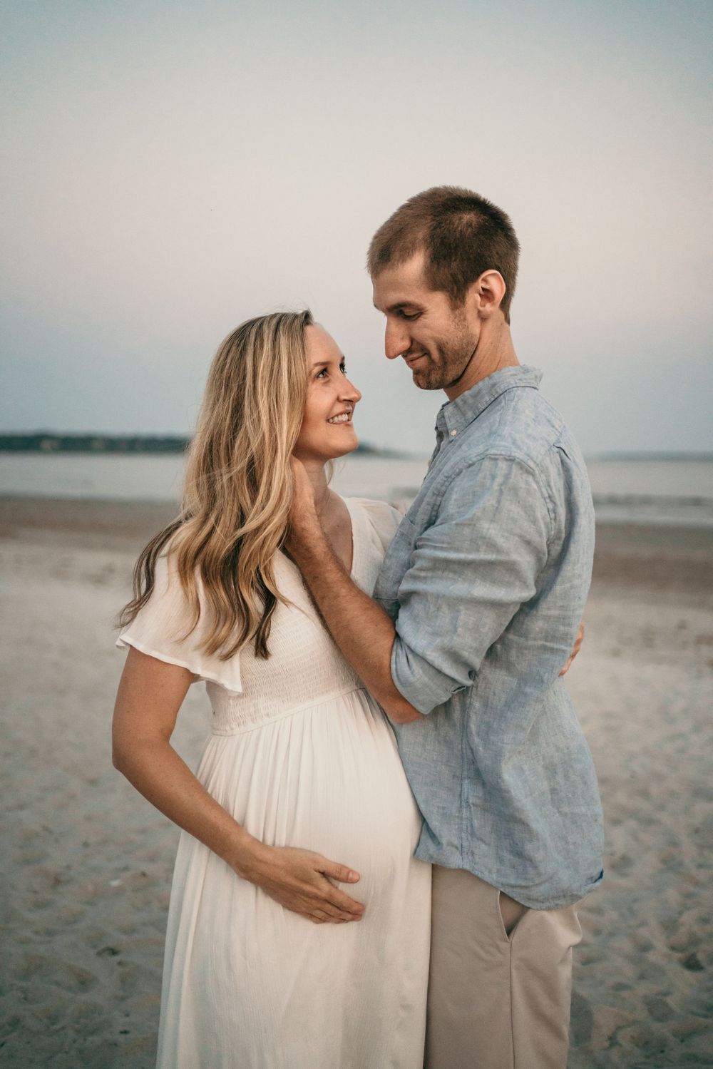 Couple embraces on beach during maternity photoshoot at dusk.