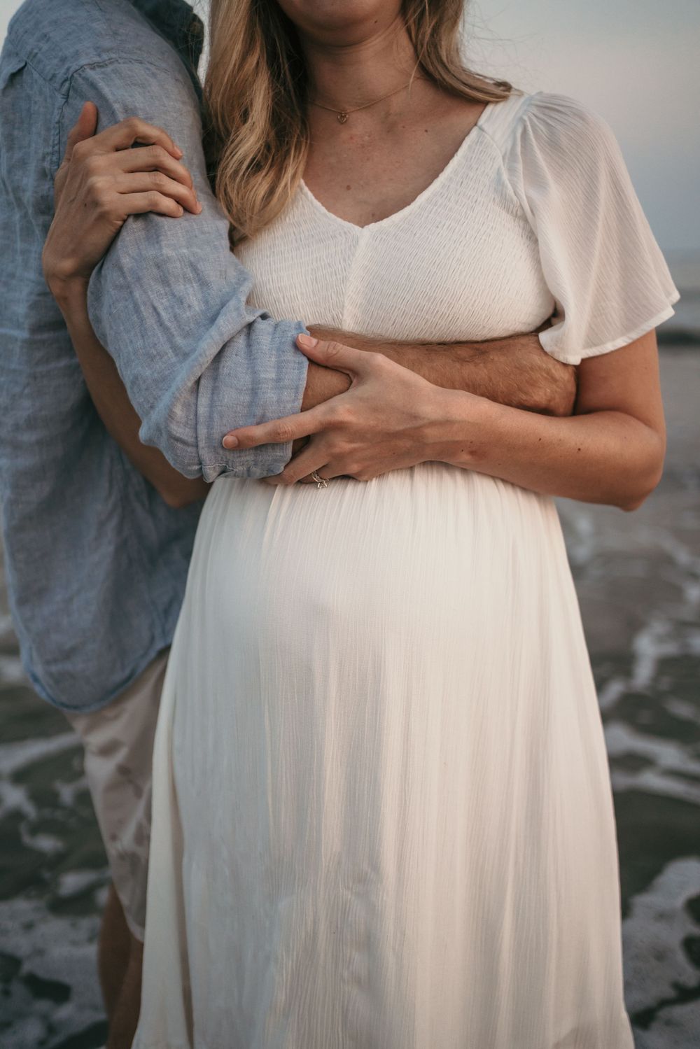 Close-up of pregnant woman in white dress holding her baby bump on beach at sunset.