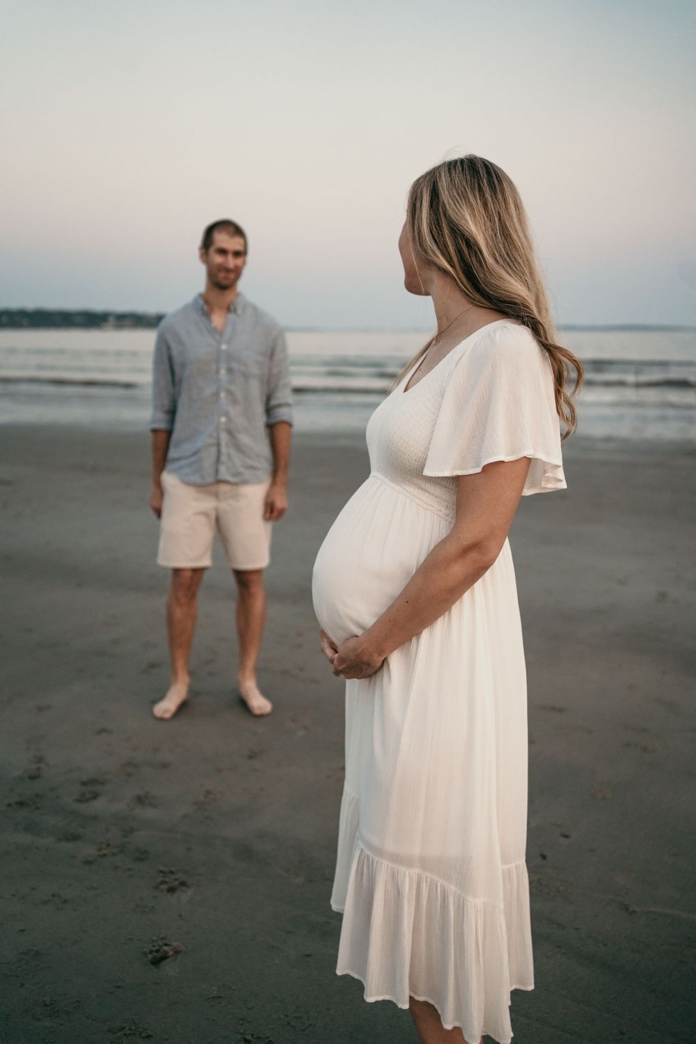 A couple stands on a beach at sunset, with woman in flowing white maternity dress.