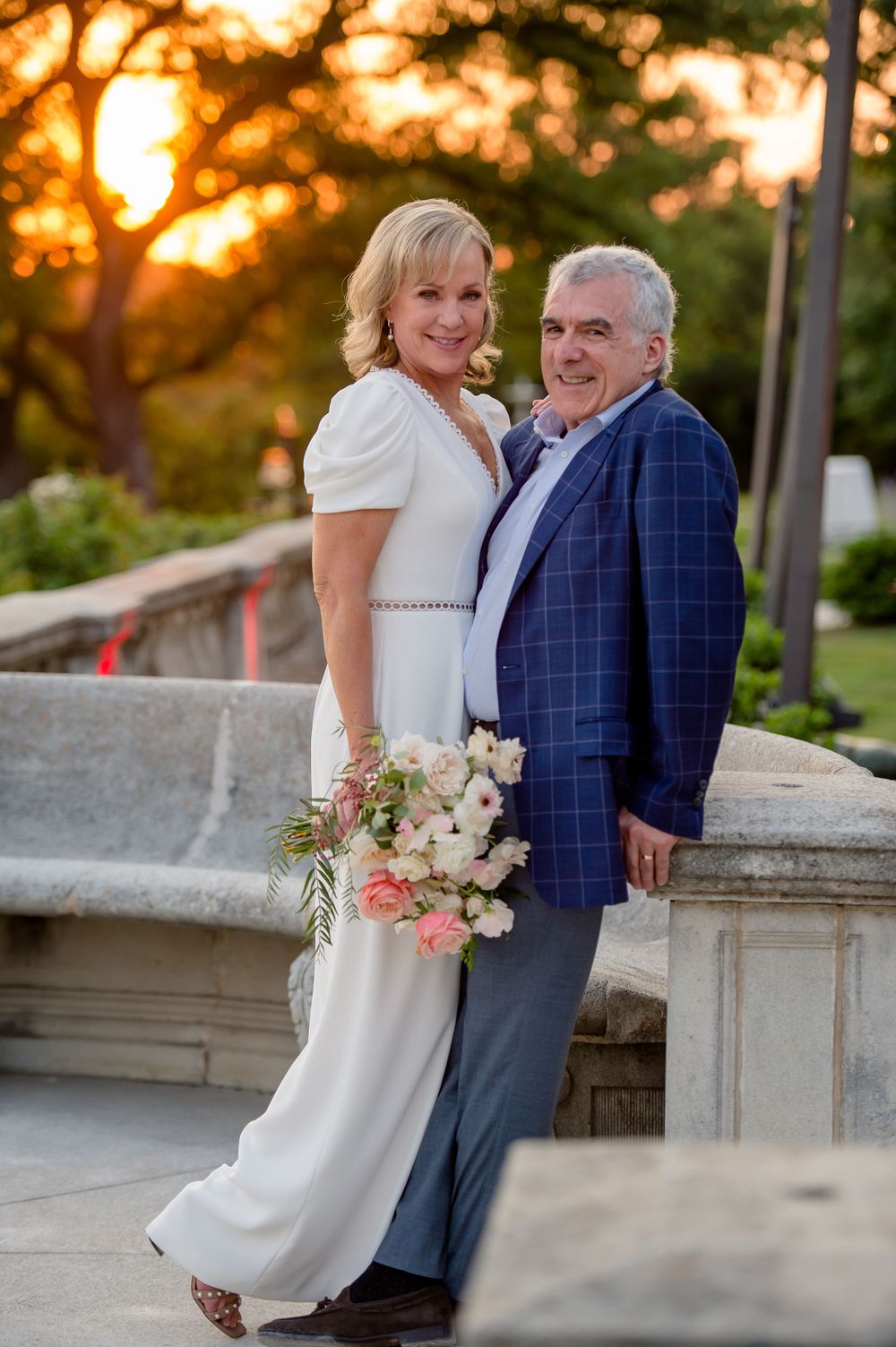 Elegant couple poses together at sunset with bride holding pink and white floral bouquet against stone railing.Commodore Perry Estate