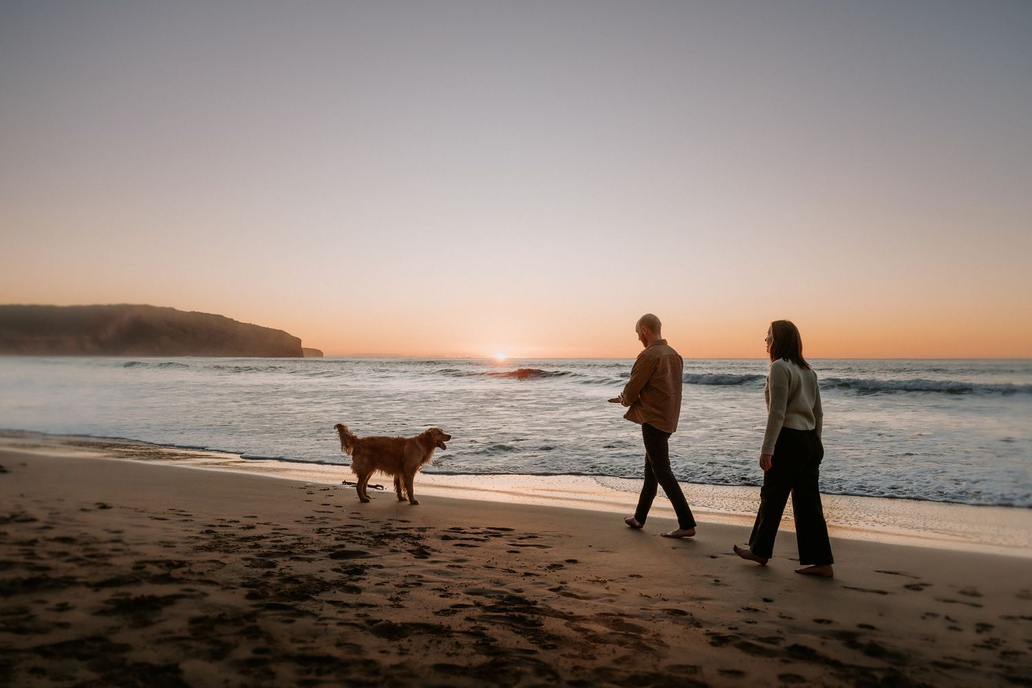 People walking their dog along a scenic beach at dusk.