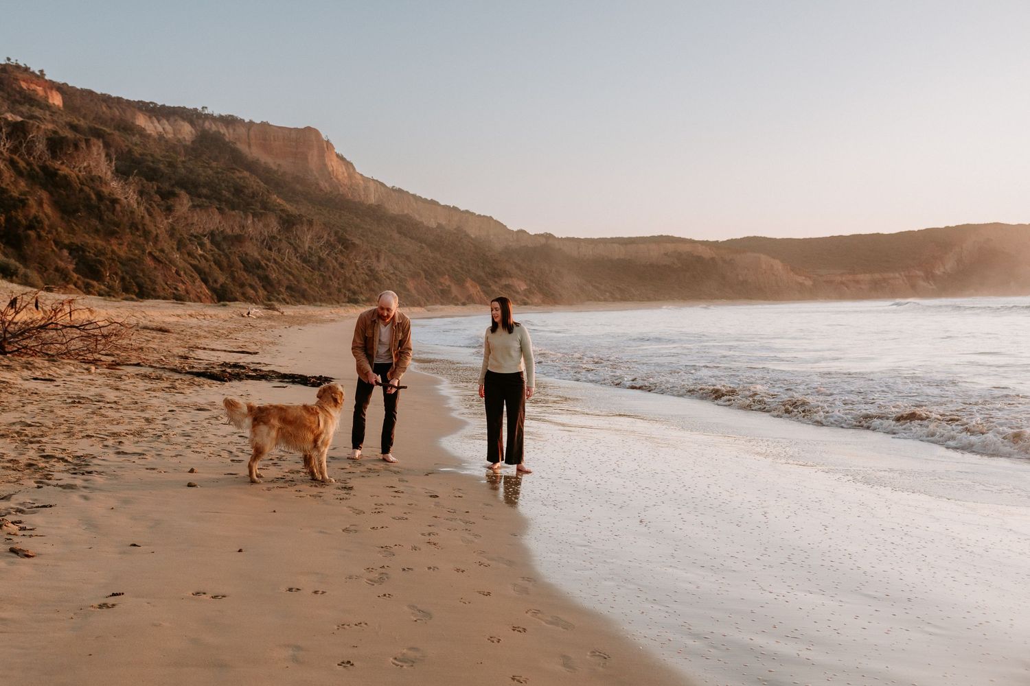 A Golden Retriever accompanies two people walking along a scenic beach at sunset with cliffs in the background.