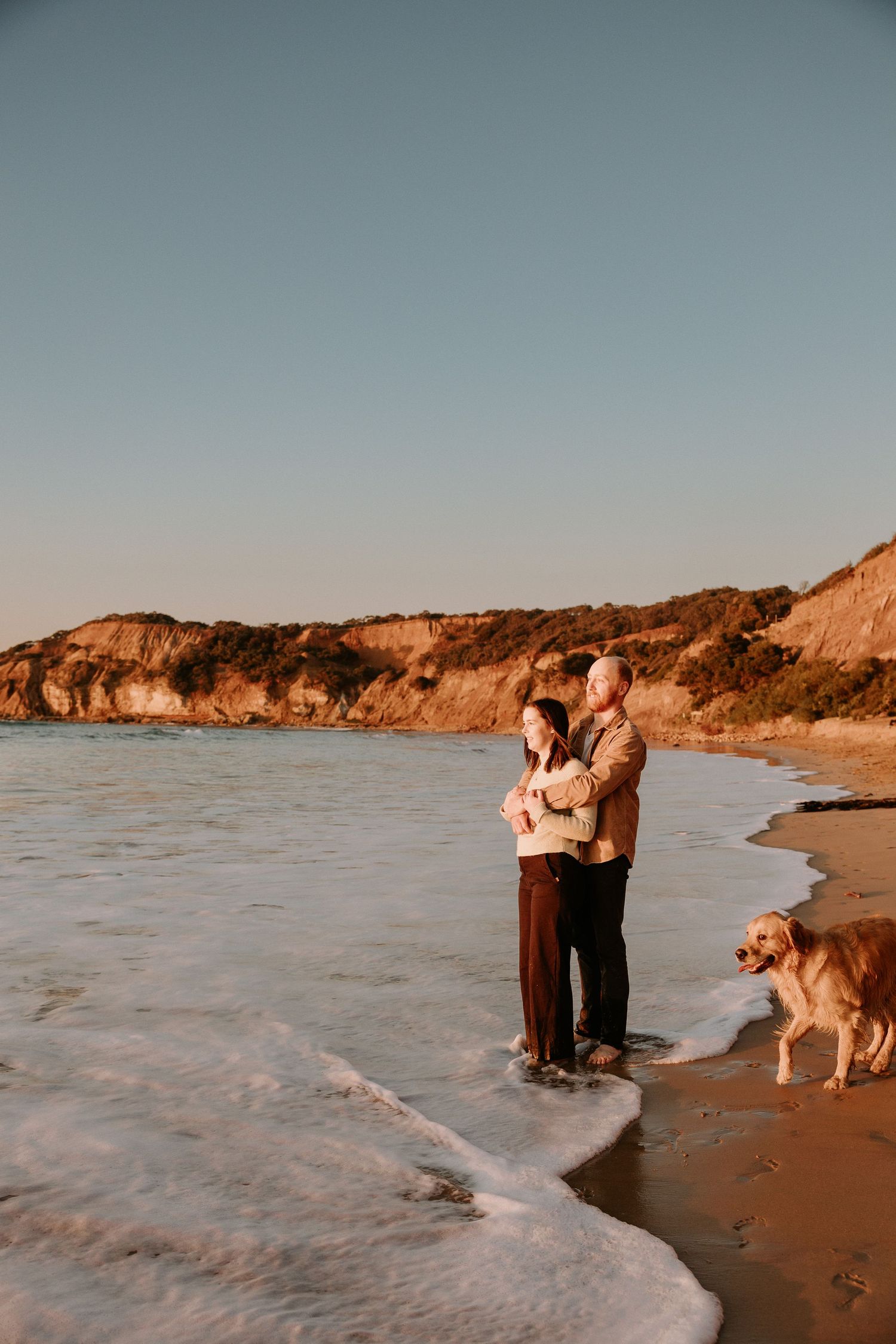 Couple embracing on a beach at sunset with their Golden Retriever standing nearby.