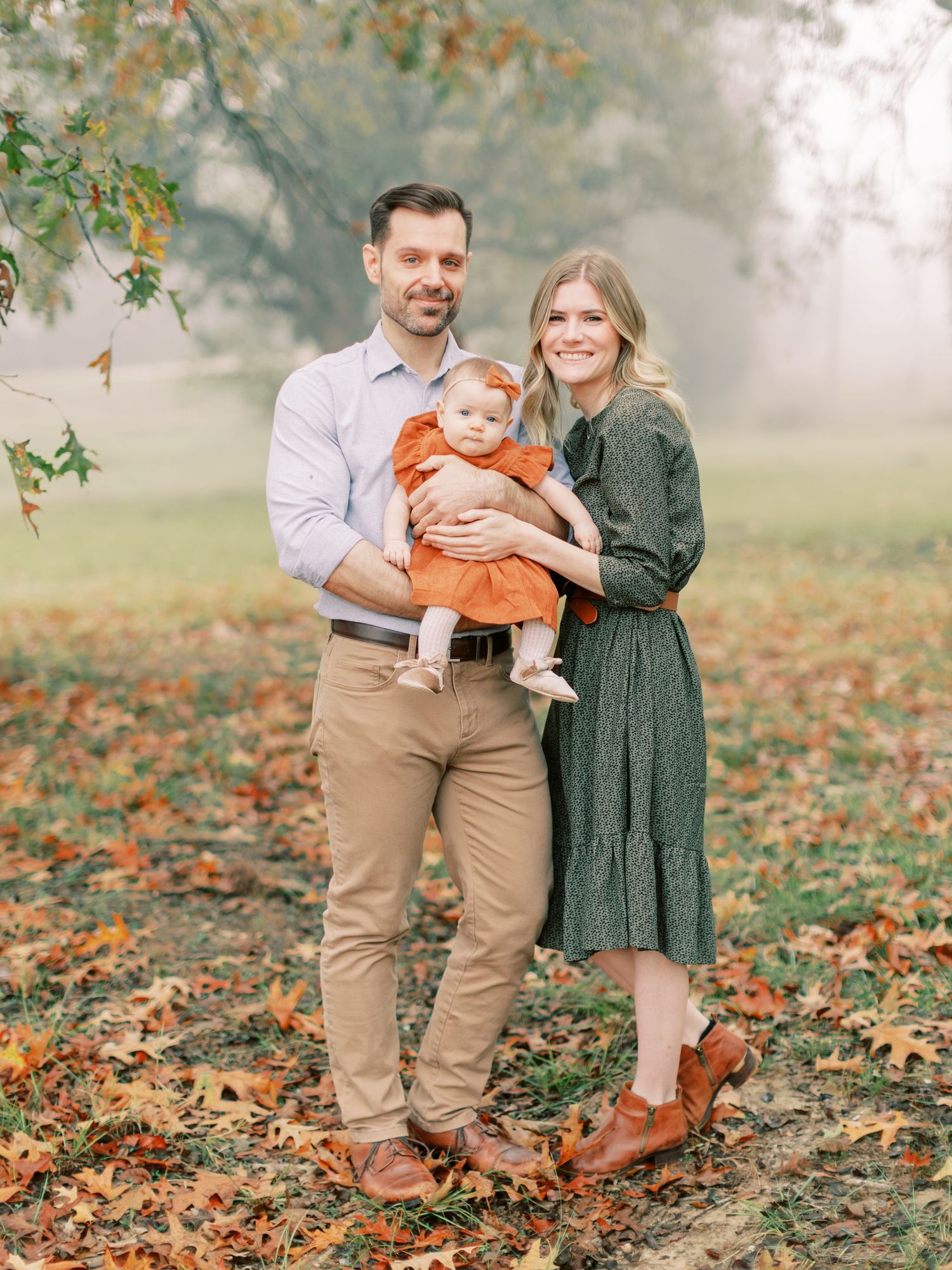 Family of three posing outdoors on autumn day with fallen leaves.