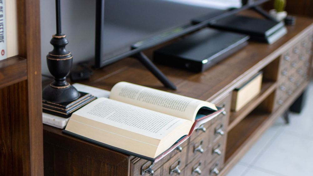 An open book displayed on a wooden shelf with a black table lamp nearby.