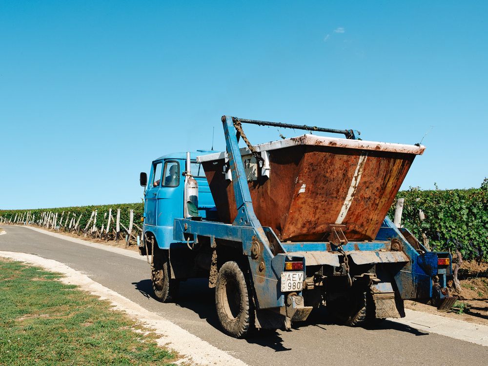 Blue industrial truck with rust-colored hopper driving through vineyard rows.
