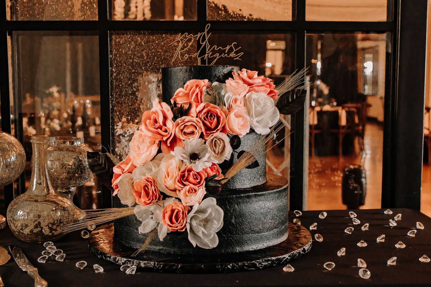 Elegant black wedding cake adorned with coral and white roses displayed on a mirrored table taken Houston Wedding Photographer.