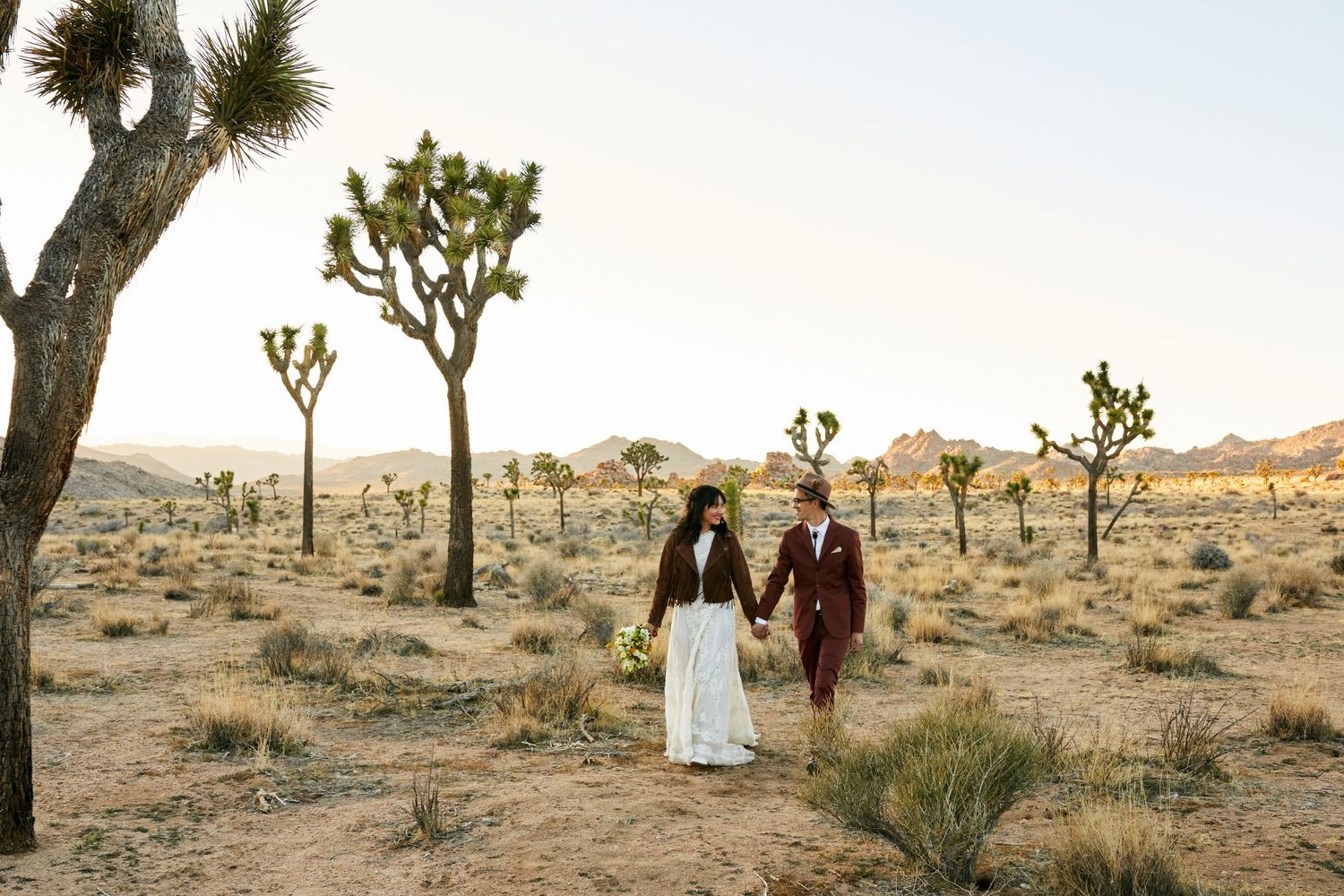 A couple walks through a desert landscape filled with Joshua trees at sunset.