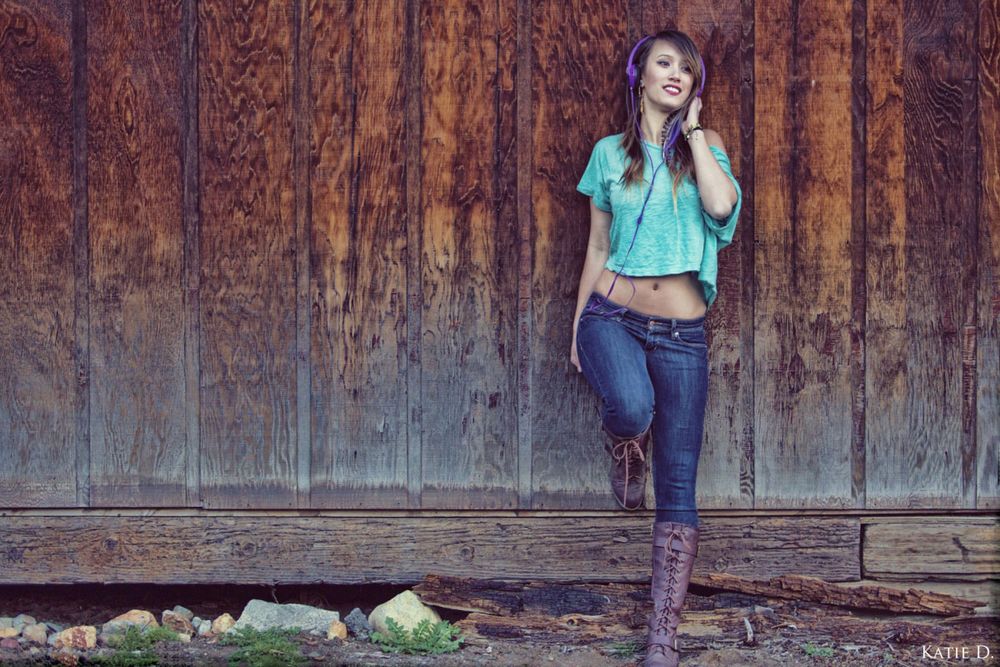 Woman in teal top and jeans leaning against a rustic wooden barn wall smiling.