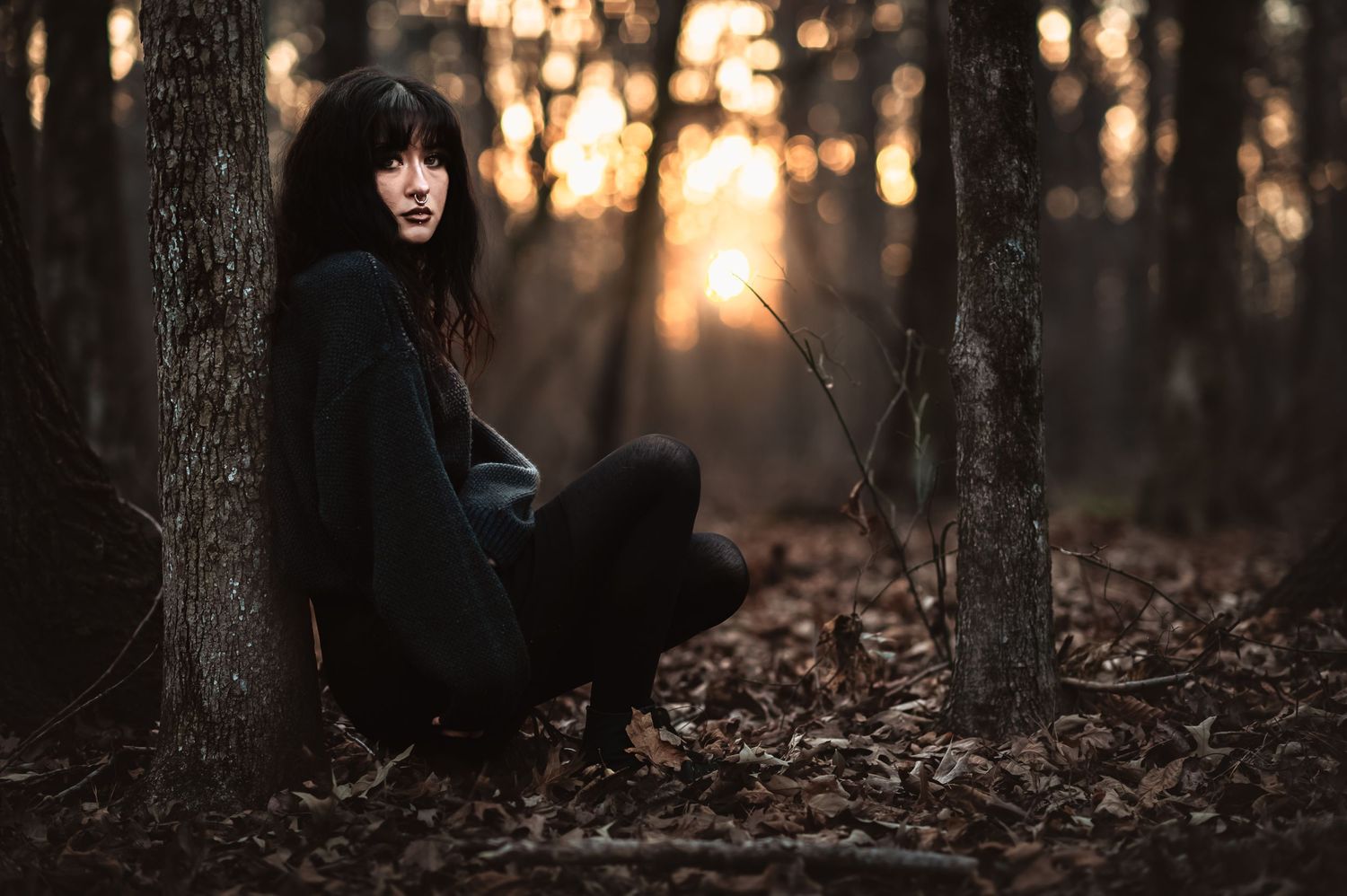 a goth high school senior leans against a tree in the woods at sunset for moody senior photos
