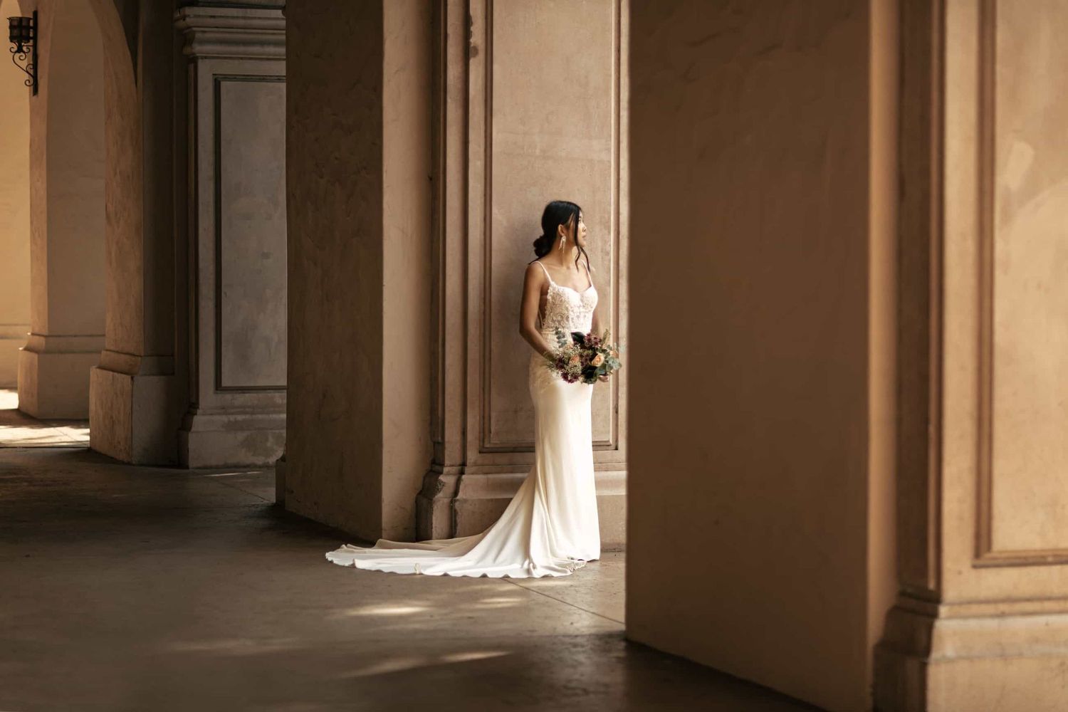 Bride in sleek white wedding gown with train stands between grand architectural columns in warm light.