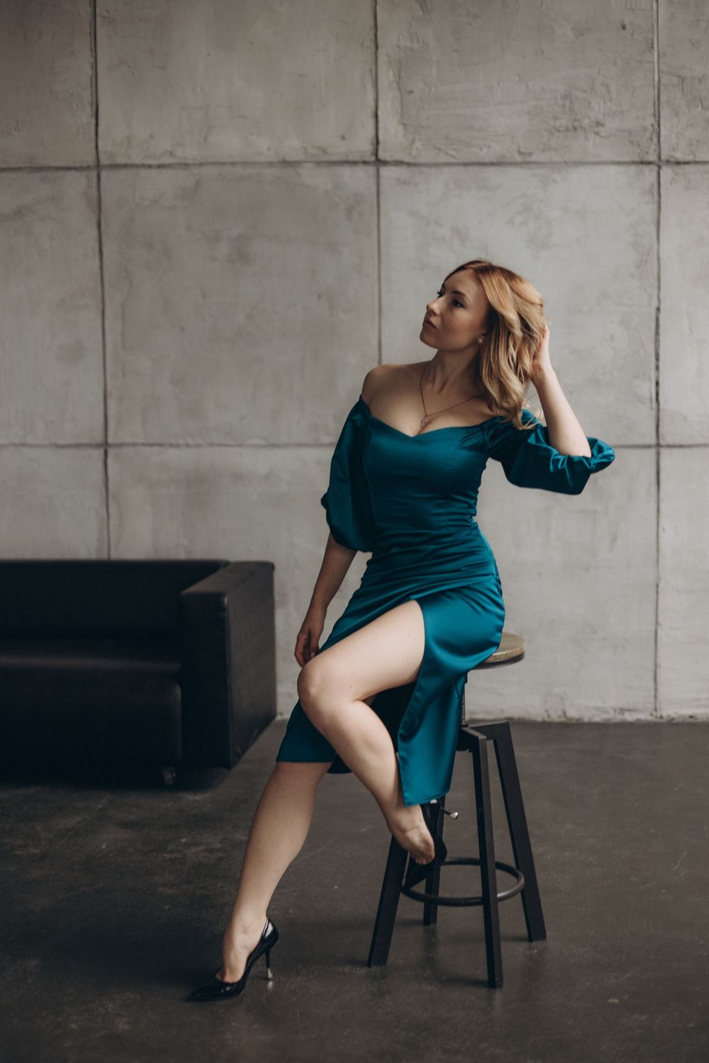 Glamorous portrait in teal dress on bar stool against concrete wall.
