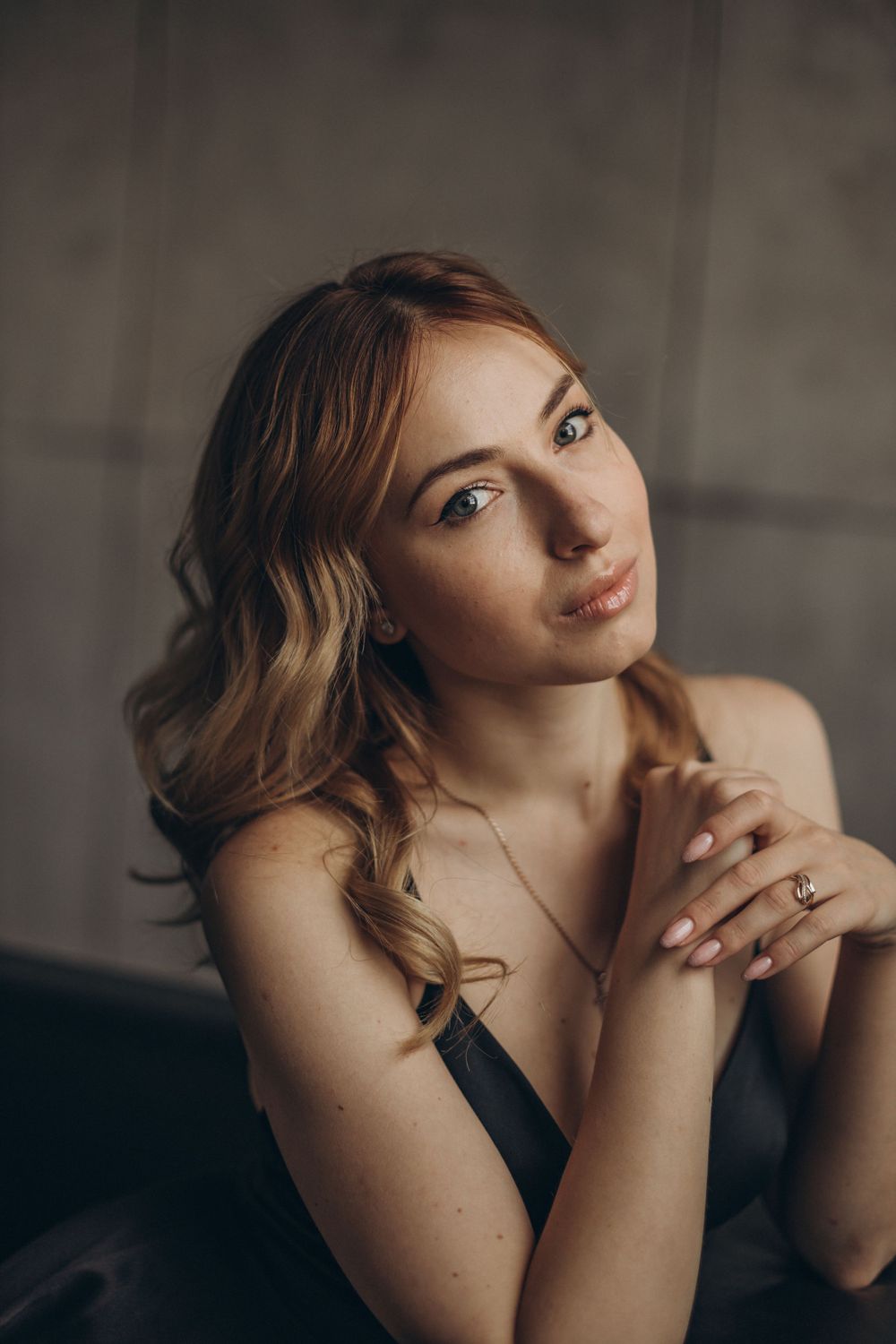 Soft portrait in muted tones featuring gentle waves of golden hair and clasped hands against a dark background.