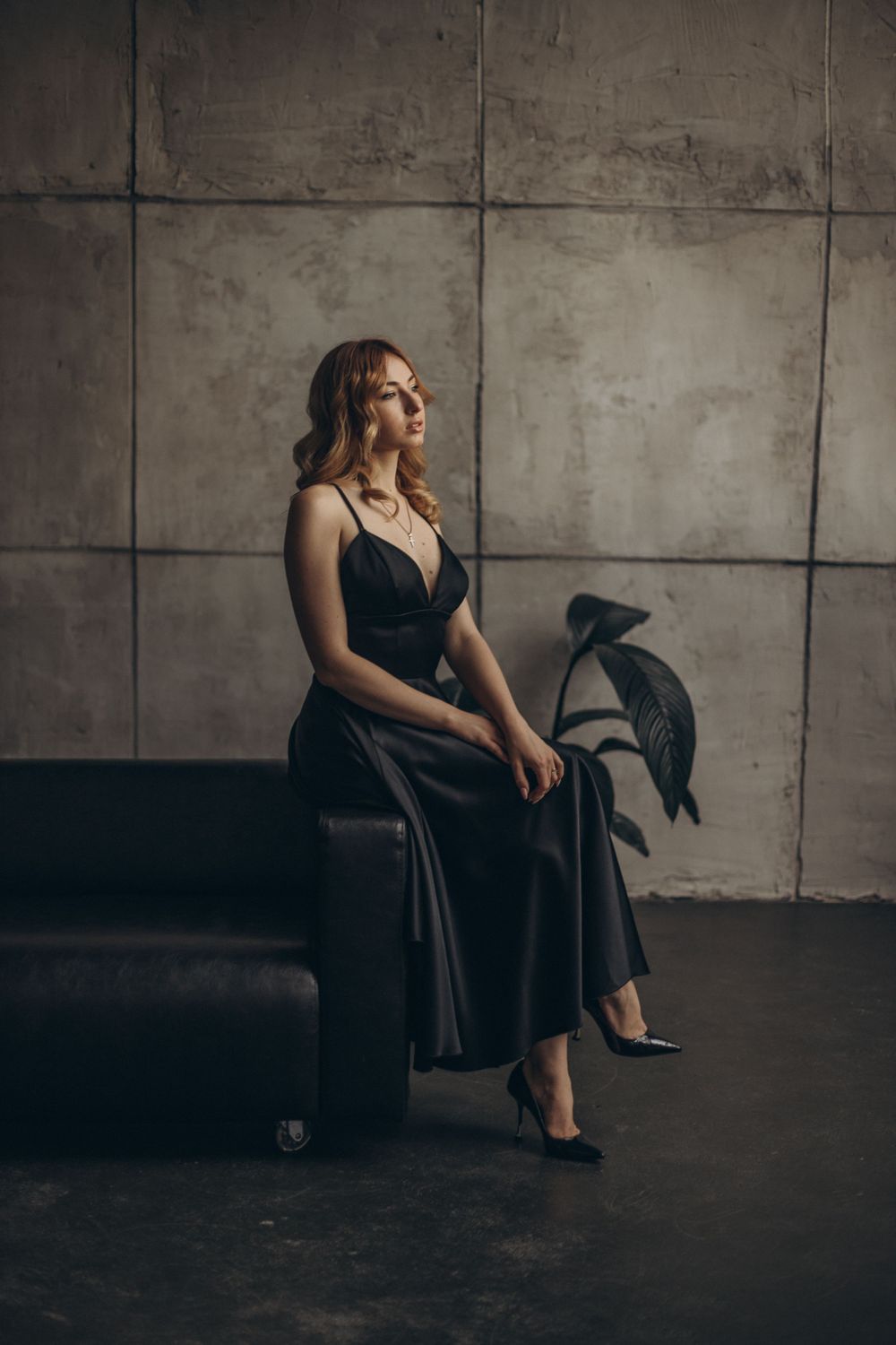 Elegant figure in a black evening gown sits gracefully on a dark couch against an industrial concrete wall.