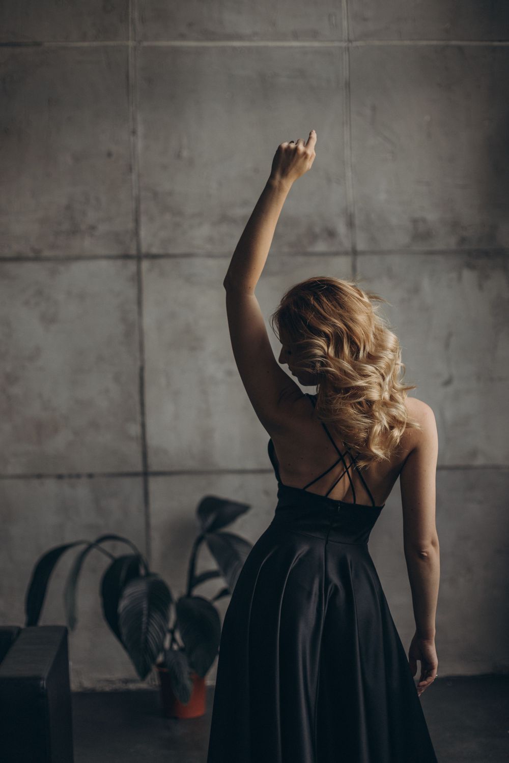 Graceful silhouette in black dress raises arm against concrete backdrop with dramatic lighting.