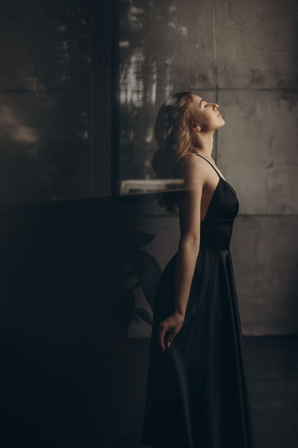 Dramatic pose in flowing black dress against moody concrete wall with artistic lighting.