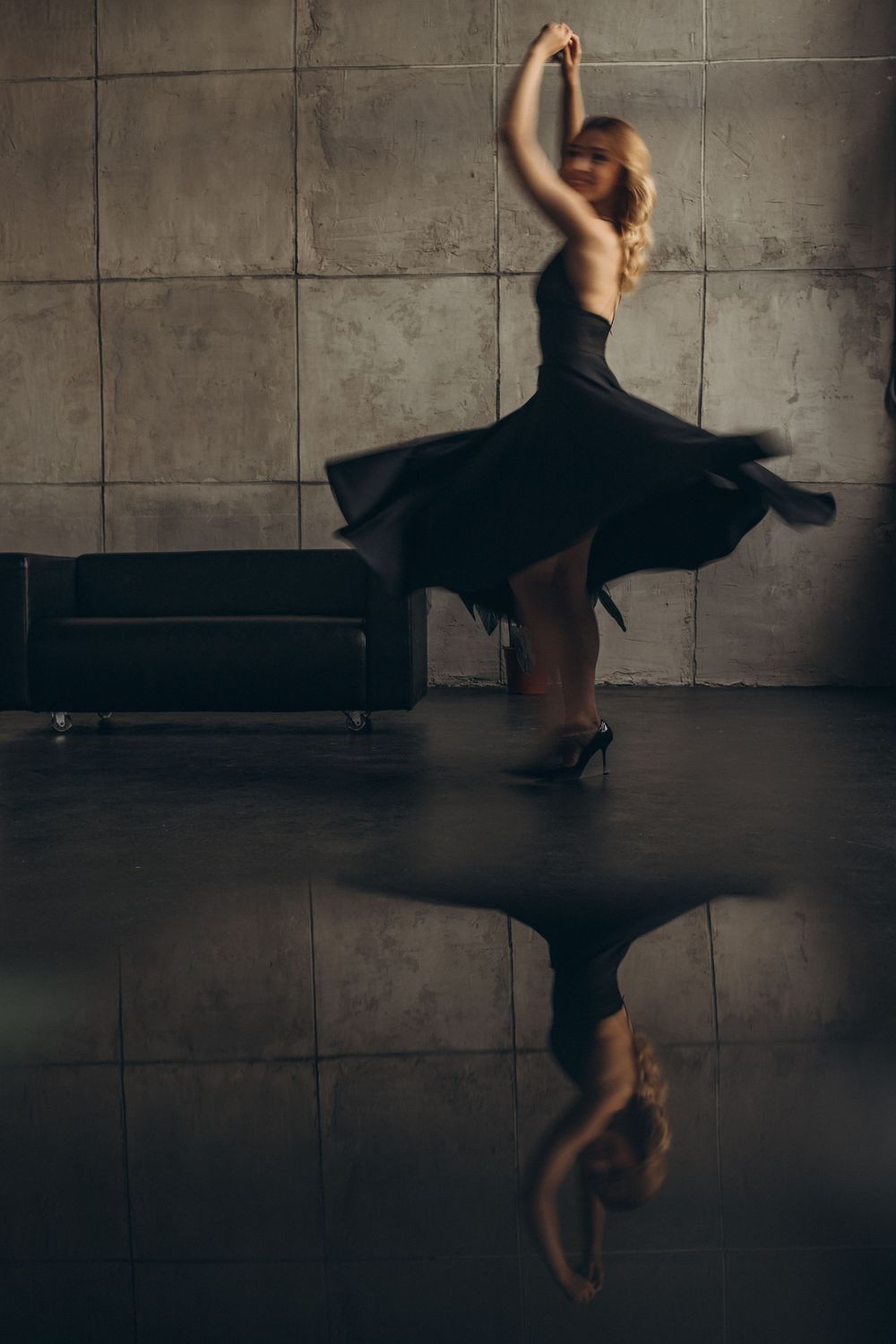 Dancing figure in black dress twirls with reflection on dark floor against concrete wall.