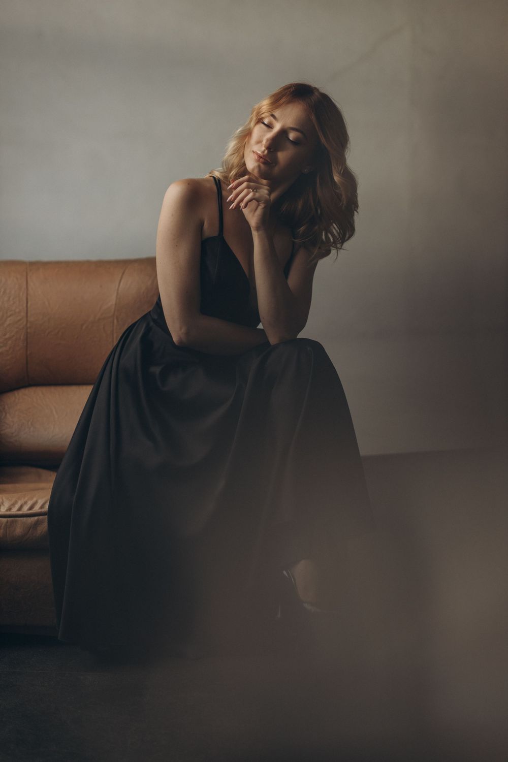 Moody portrait in black dress seated on leather couch with atmospheric lighting.