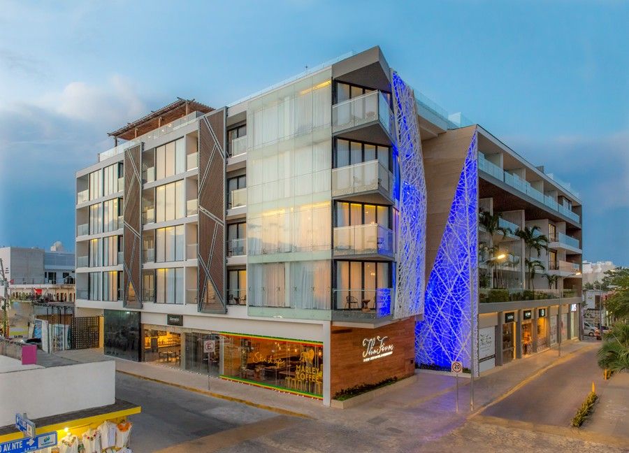 Modern apartment complex with illuminated blue architectural feature and ground-floor retail shops at dusk.