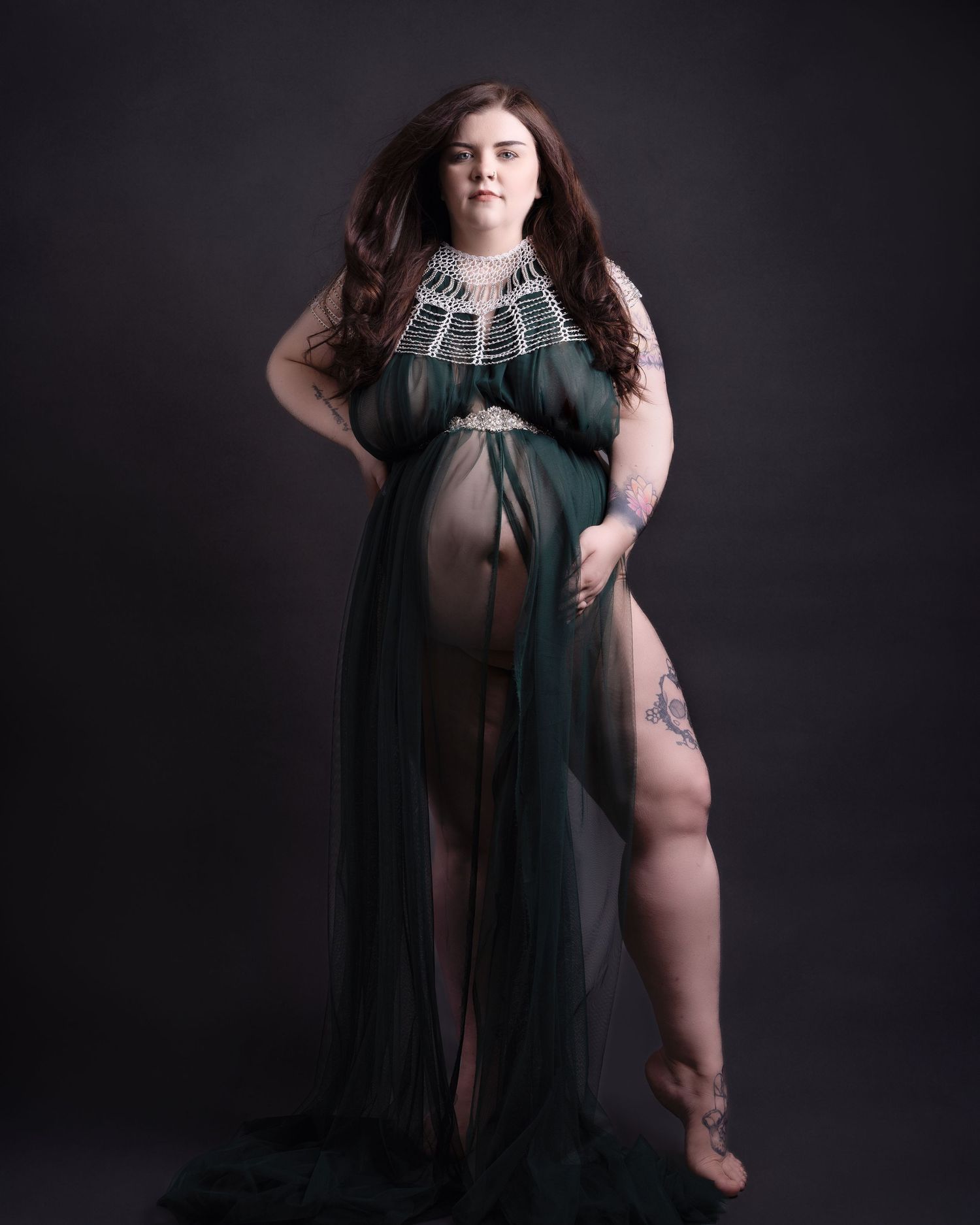 Maternity portrait in sheer green gown showing silhouette against dark backdrop.