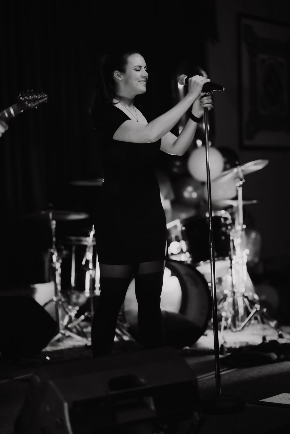 Singer in black dress performing on stage with microphone and drum kit visible in background.