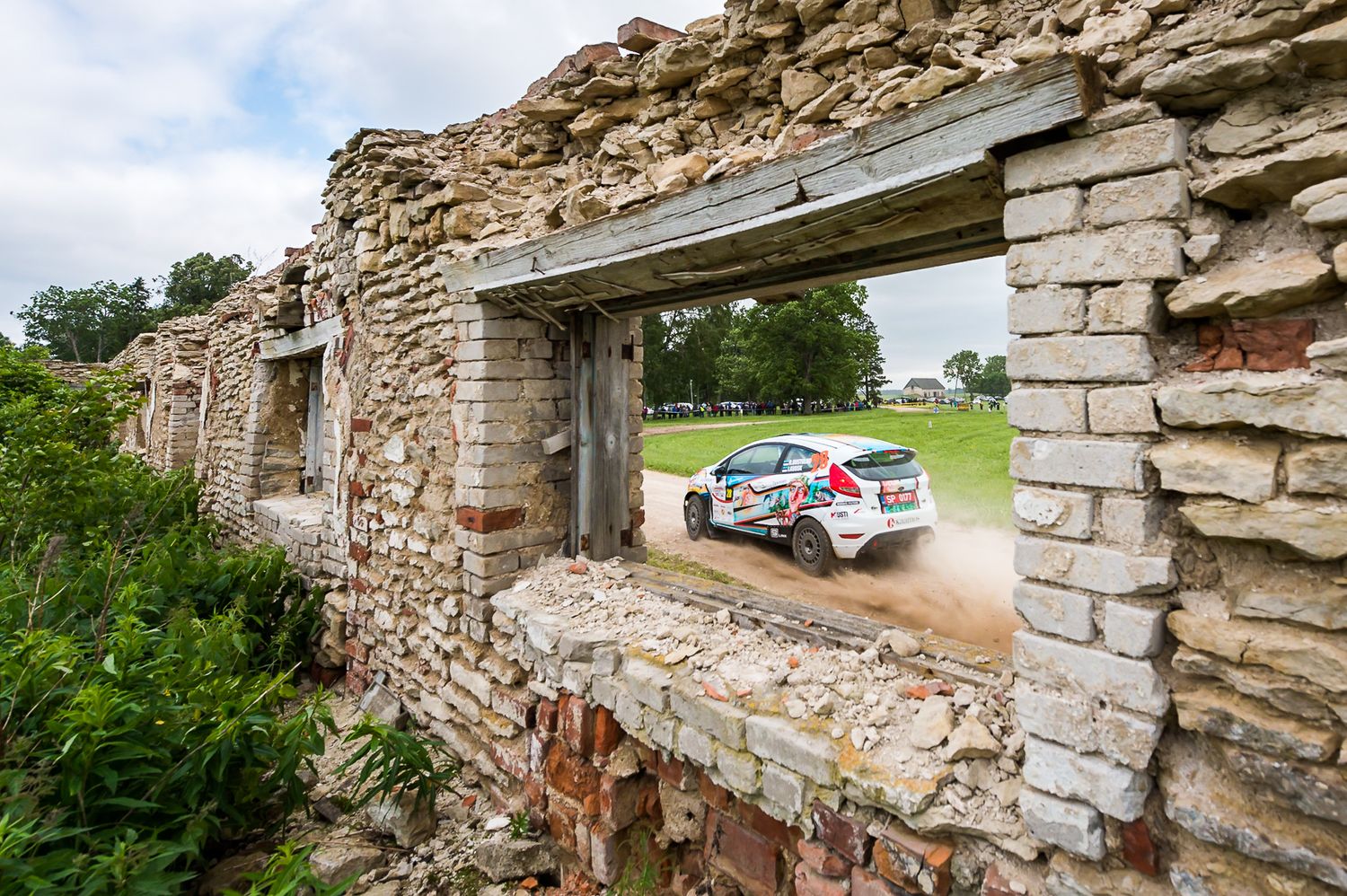 Rally car speeds past old stone ruins with windows framing the action.