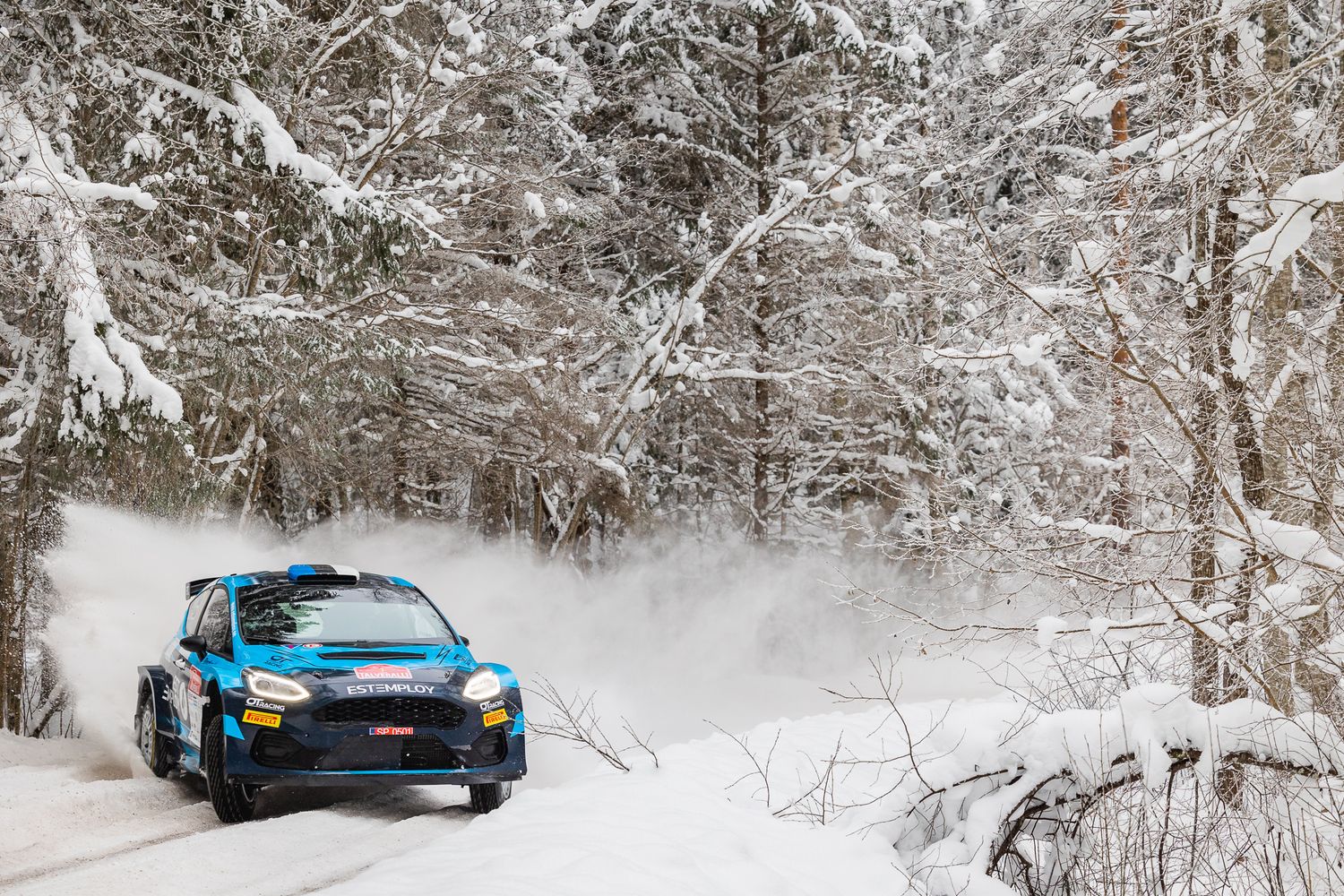 Rally car racing through snow-covered forest trail creating powder spray.