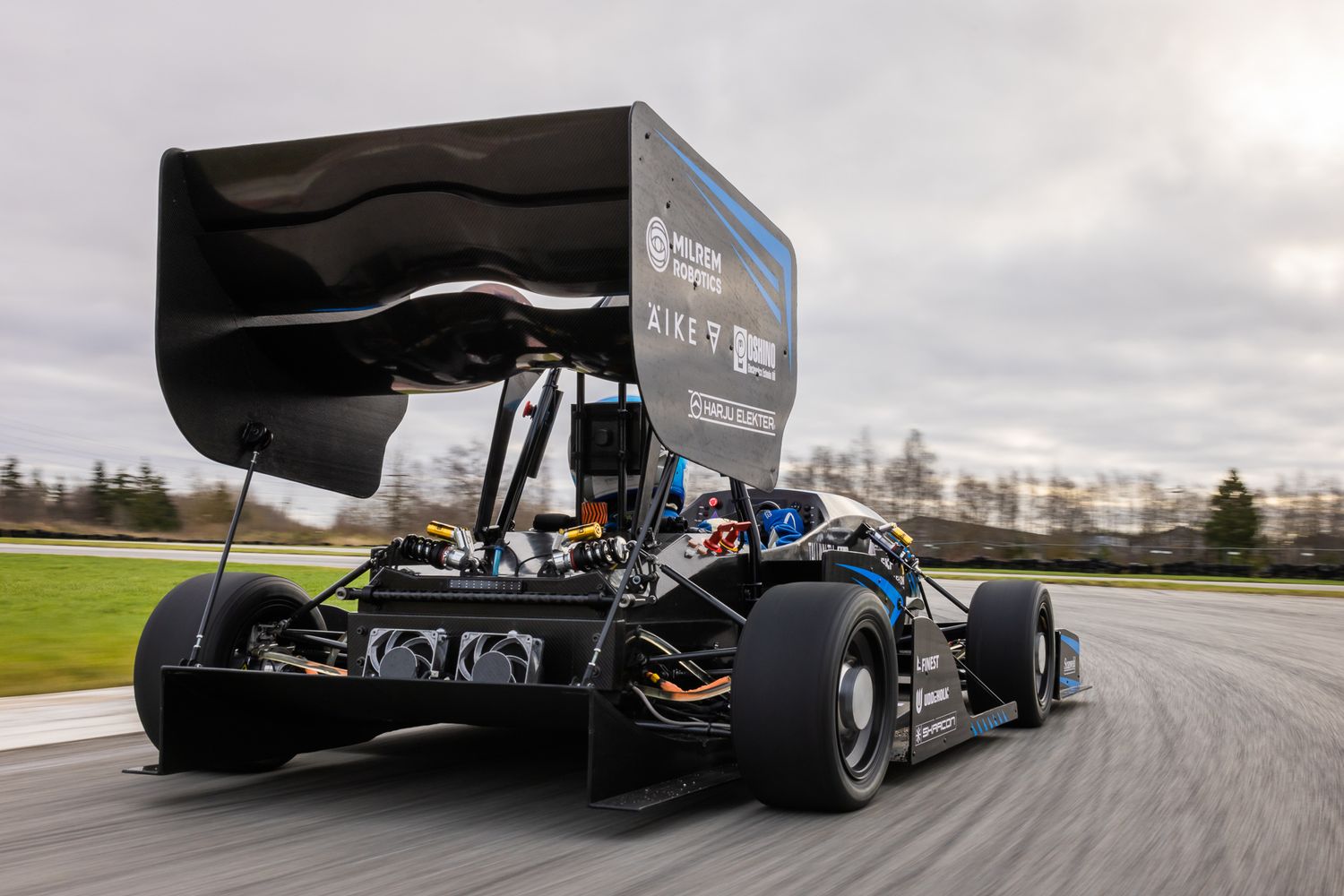 Black racing car with large rear wing on a wet racetrack under cloudy skies.