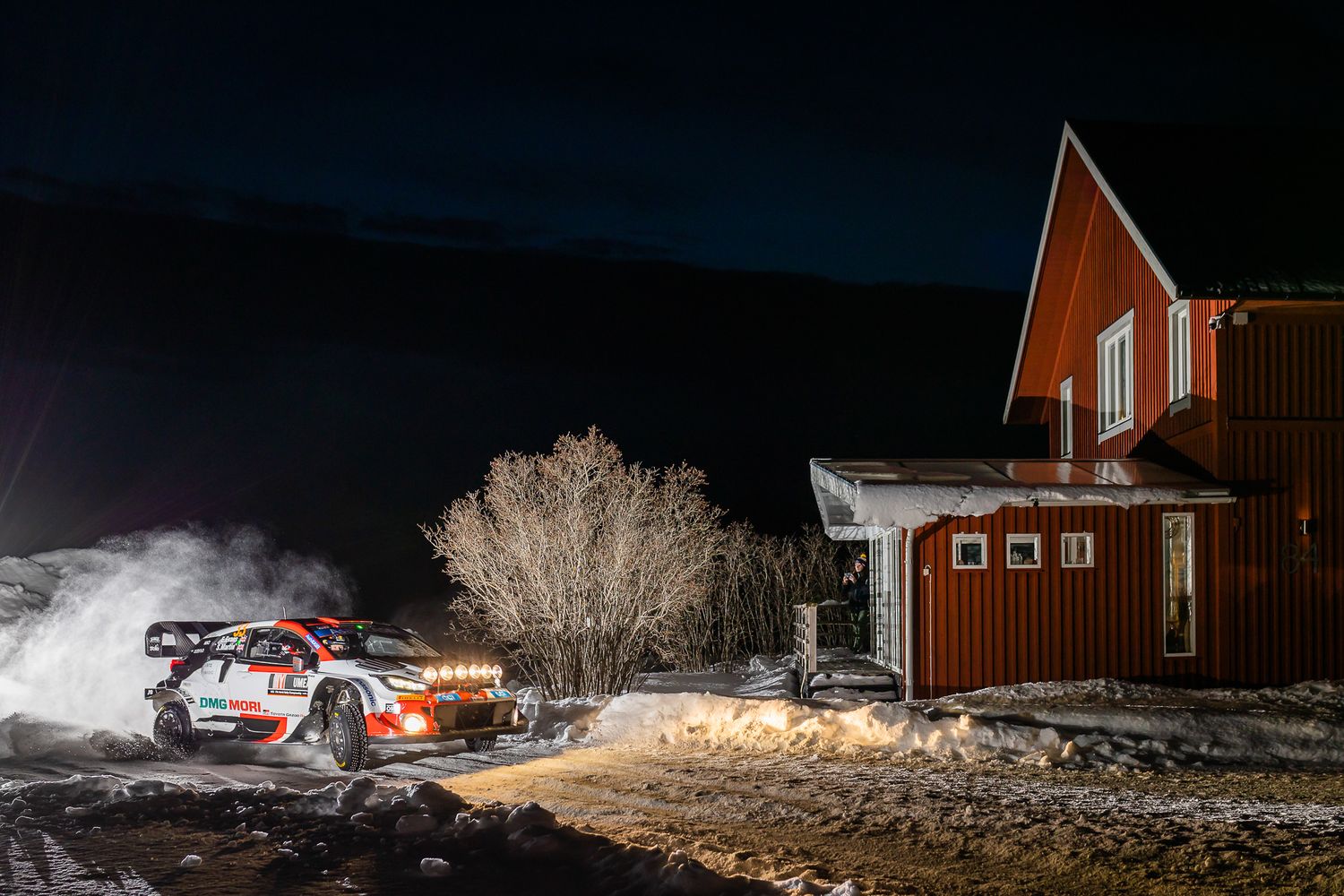 Rally car parked next to red wooden buildings in snowy winter nighttime scene.