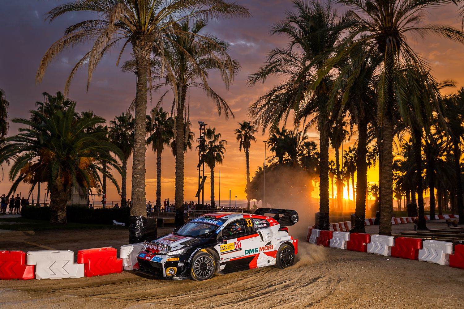 Rally car races through palm-lined course at sunset with dramatic sky and orange lighting.