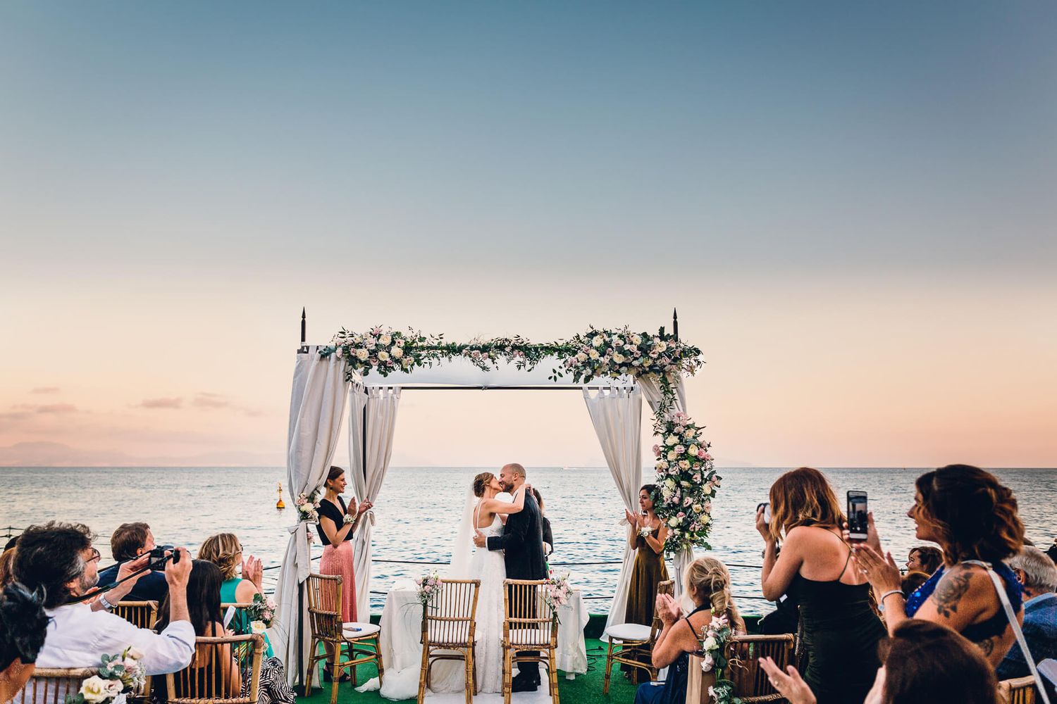 Oceanside wedding ceremony setup with floral arch and white drapes against sunset sky.