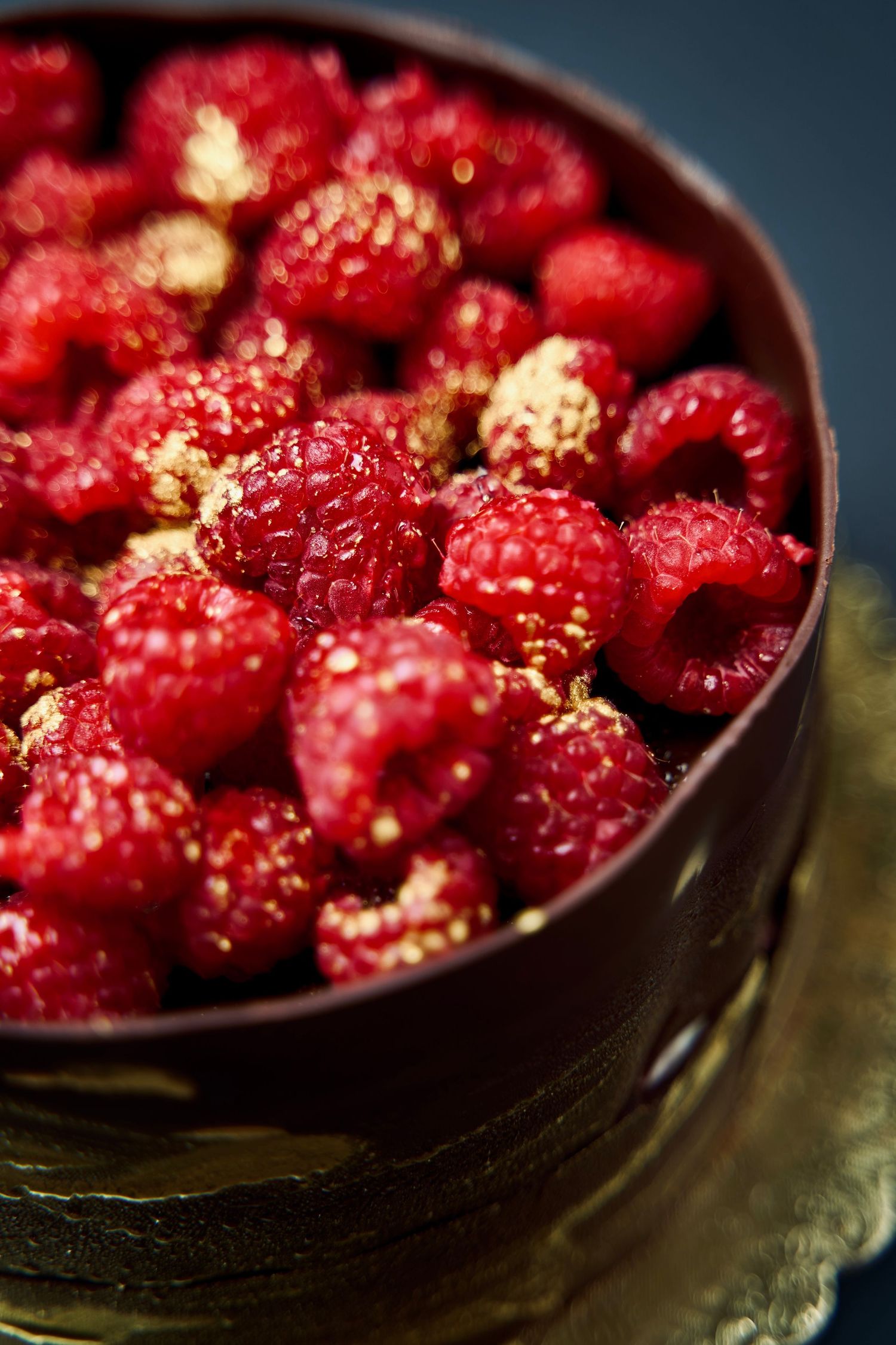 Fresh red raspberries dusted with gold in a dark ceramic bowl.