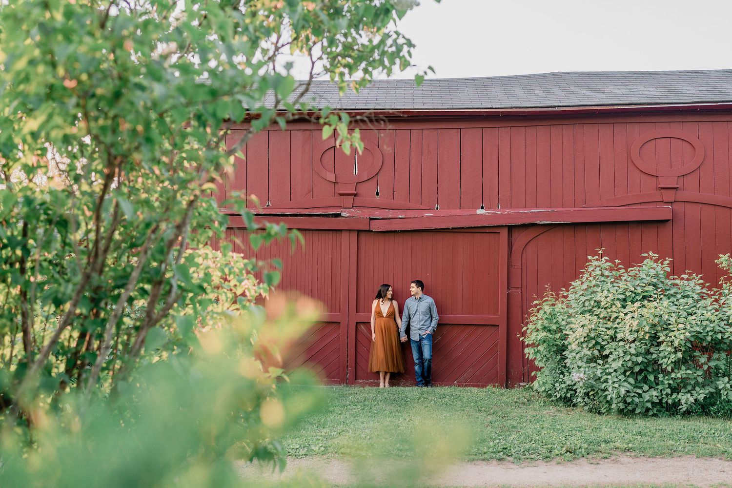 couple holding hands and leaning on old rustic red barn for engagement photos in Southern Maine