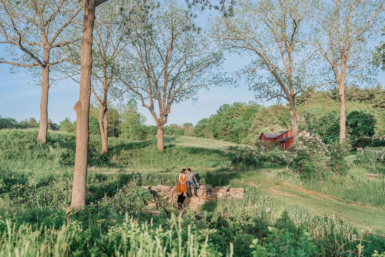 couple sitting on stone bridge surrounded by trees during engagement session at hamilton house in Maine