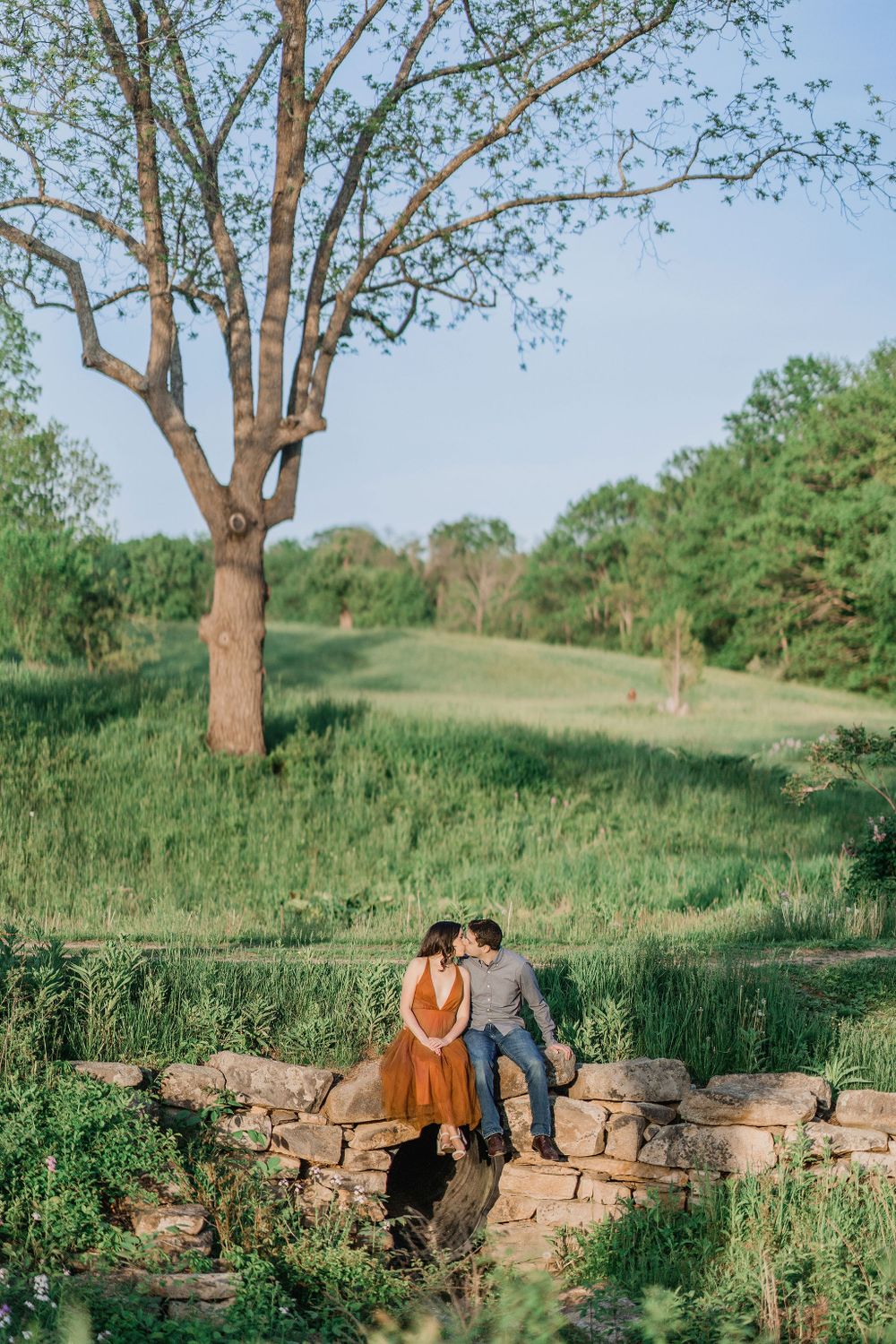 woman in orange dress kissing man sitting on stone arch bridge during Maine engagement session