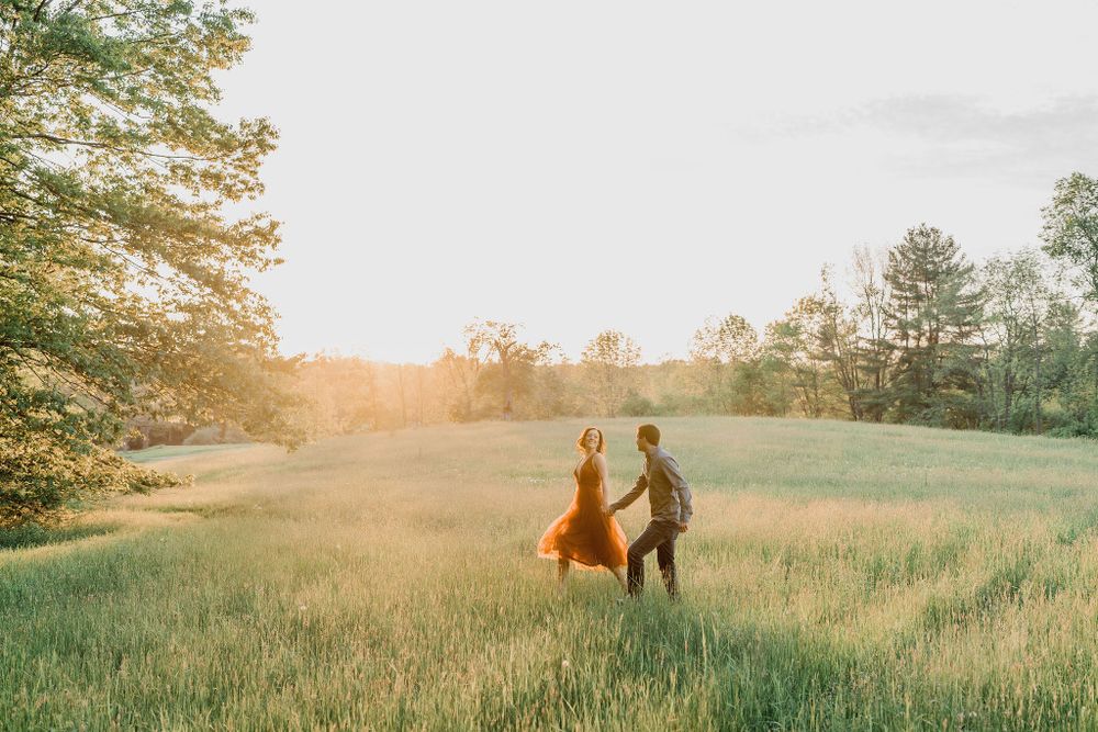 woman in rust dress holding hands with man and running in long grass during golden hour